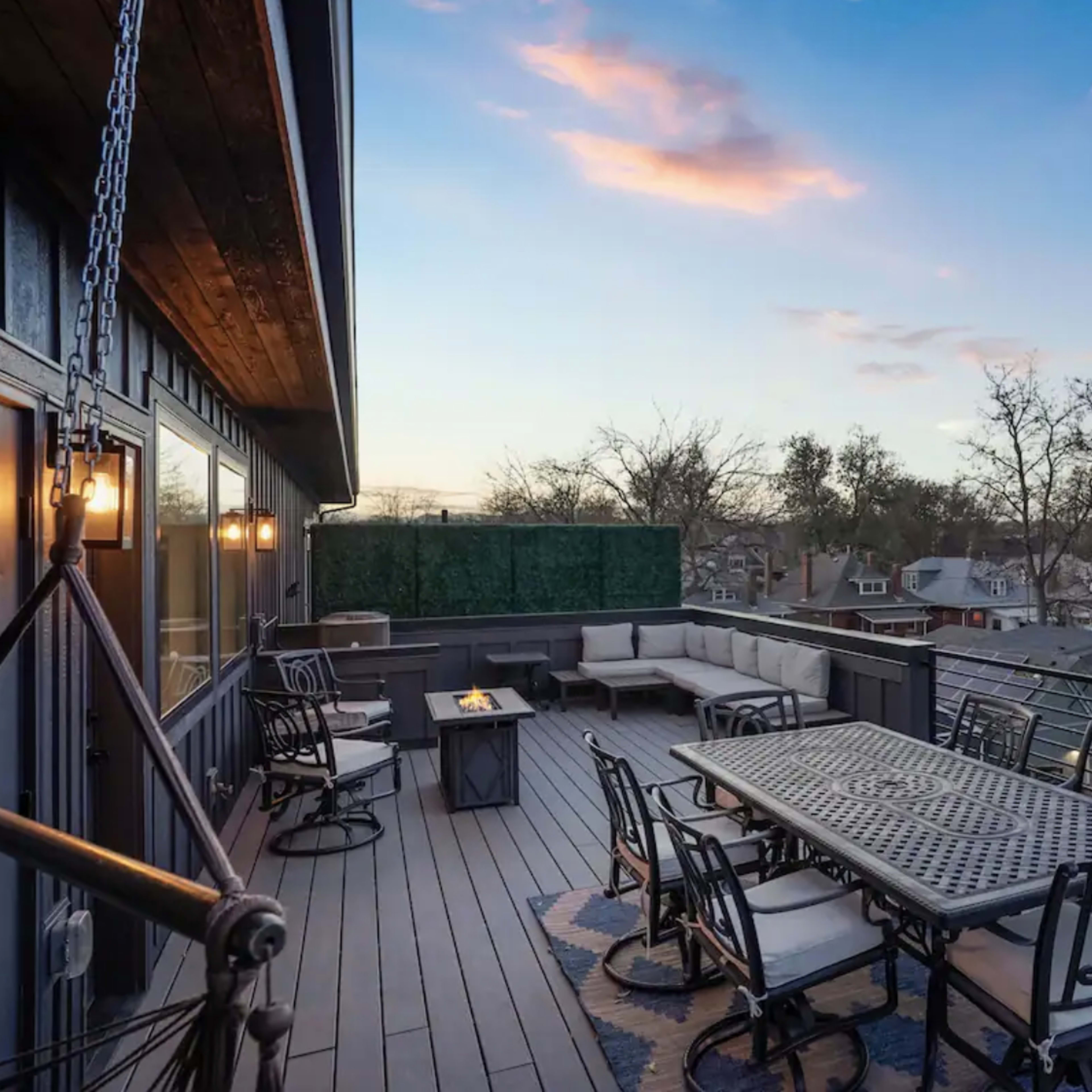 A spacious rooftop deck features a dining table, seating area, and views of trees and rooftops under a pastel sky.