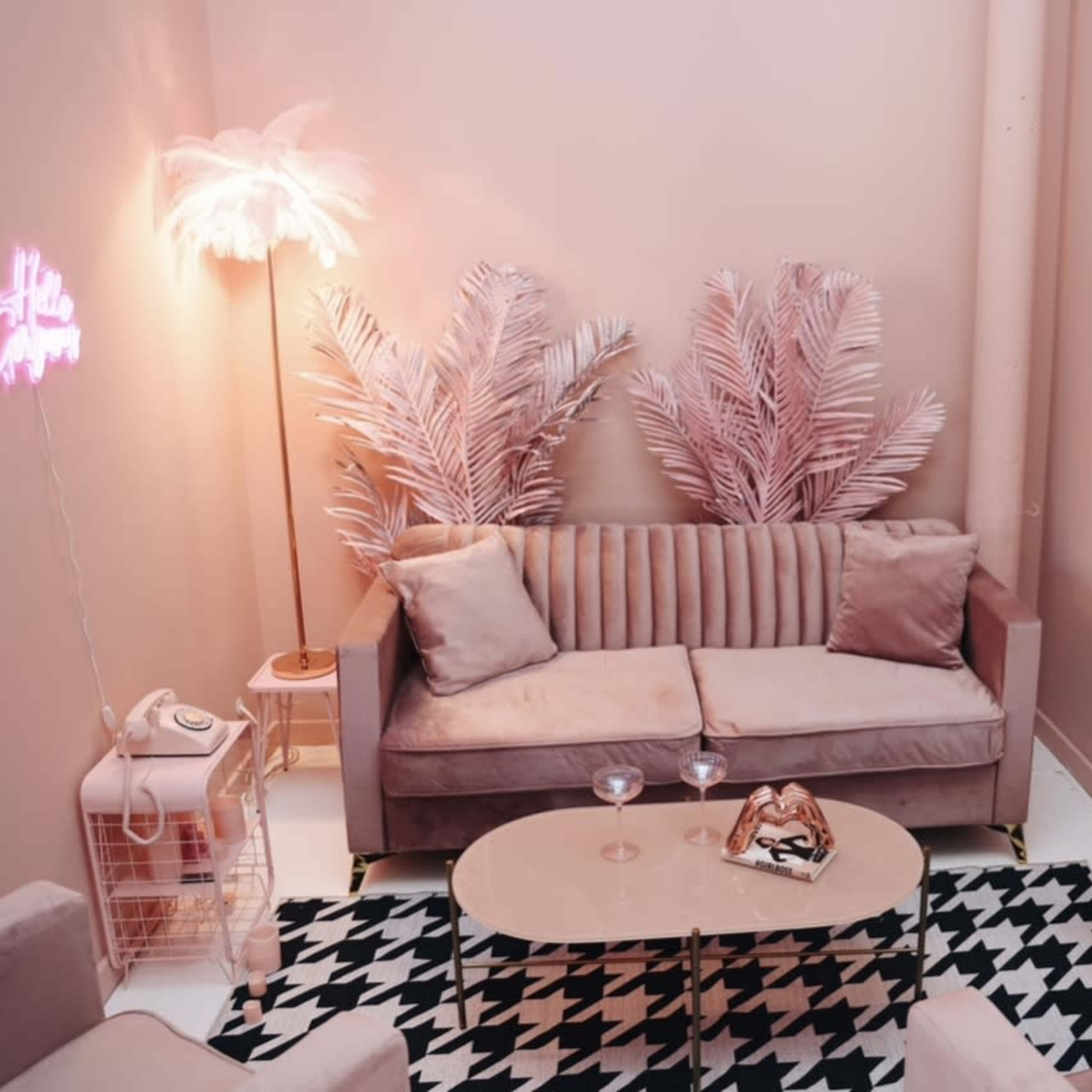 The scene features a cozy pink-themed lounge area with a sofa, patterned rug, and decorative plants.