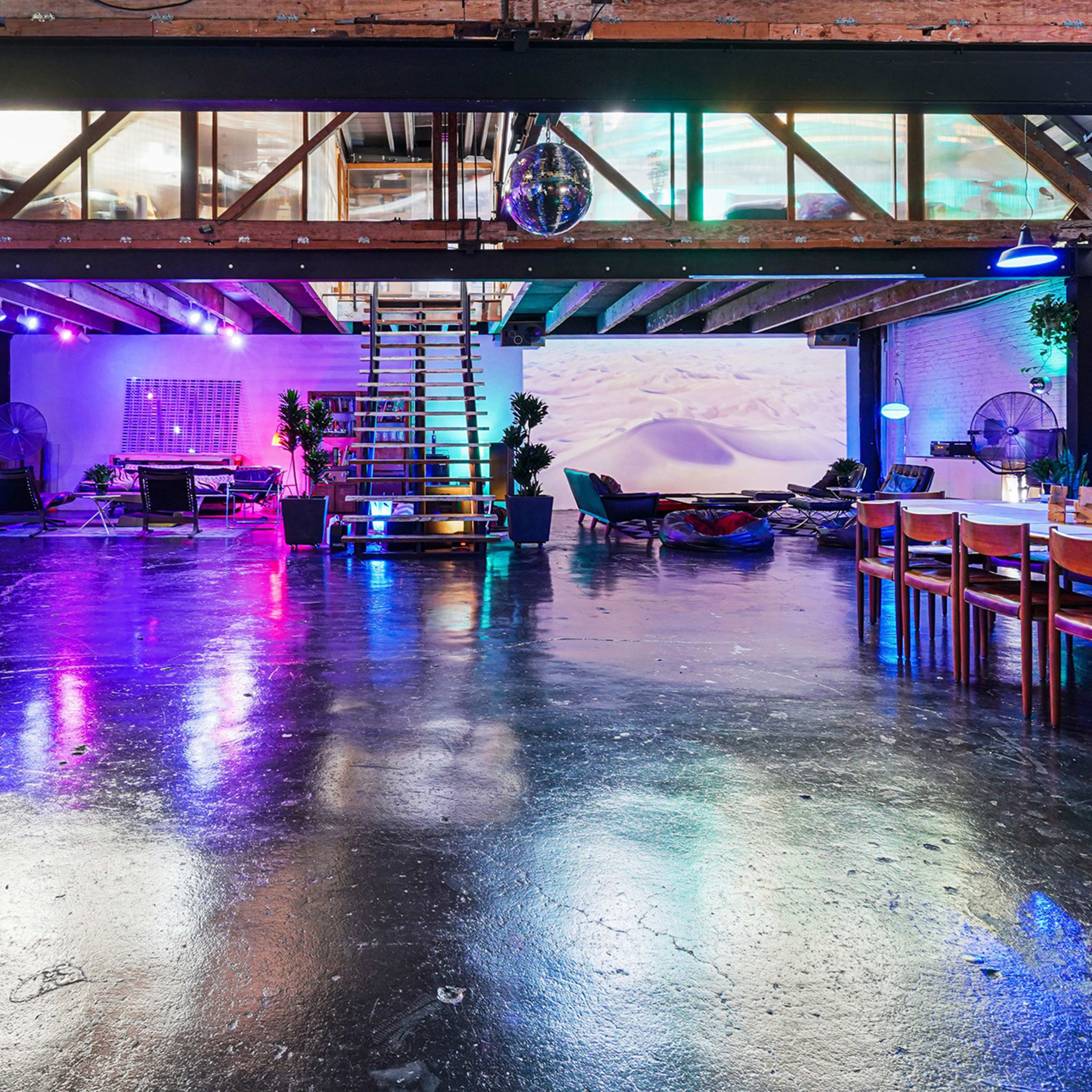 A spacious, modern interior with colorful ambient lighting, exposed beams, and a mix of seating areas featuring a dining table, lounge chairs, and a staircase.