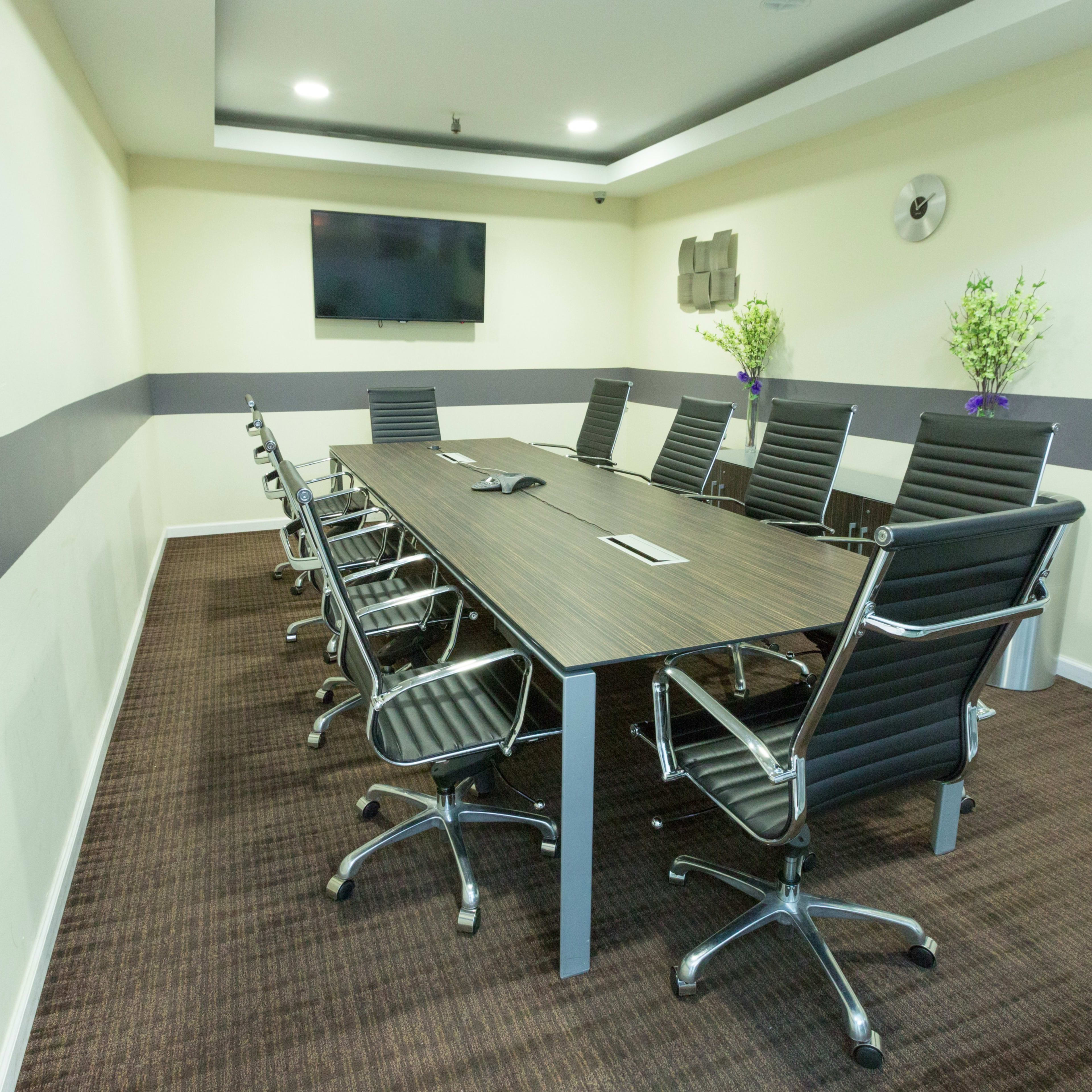 A modern conference room with a long rectangular table surrounded by black rolling chairs and a wall-mounted television.