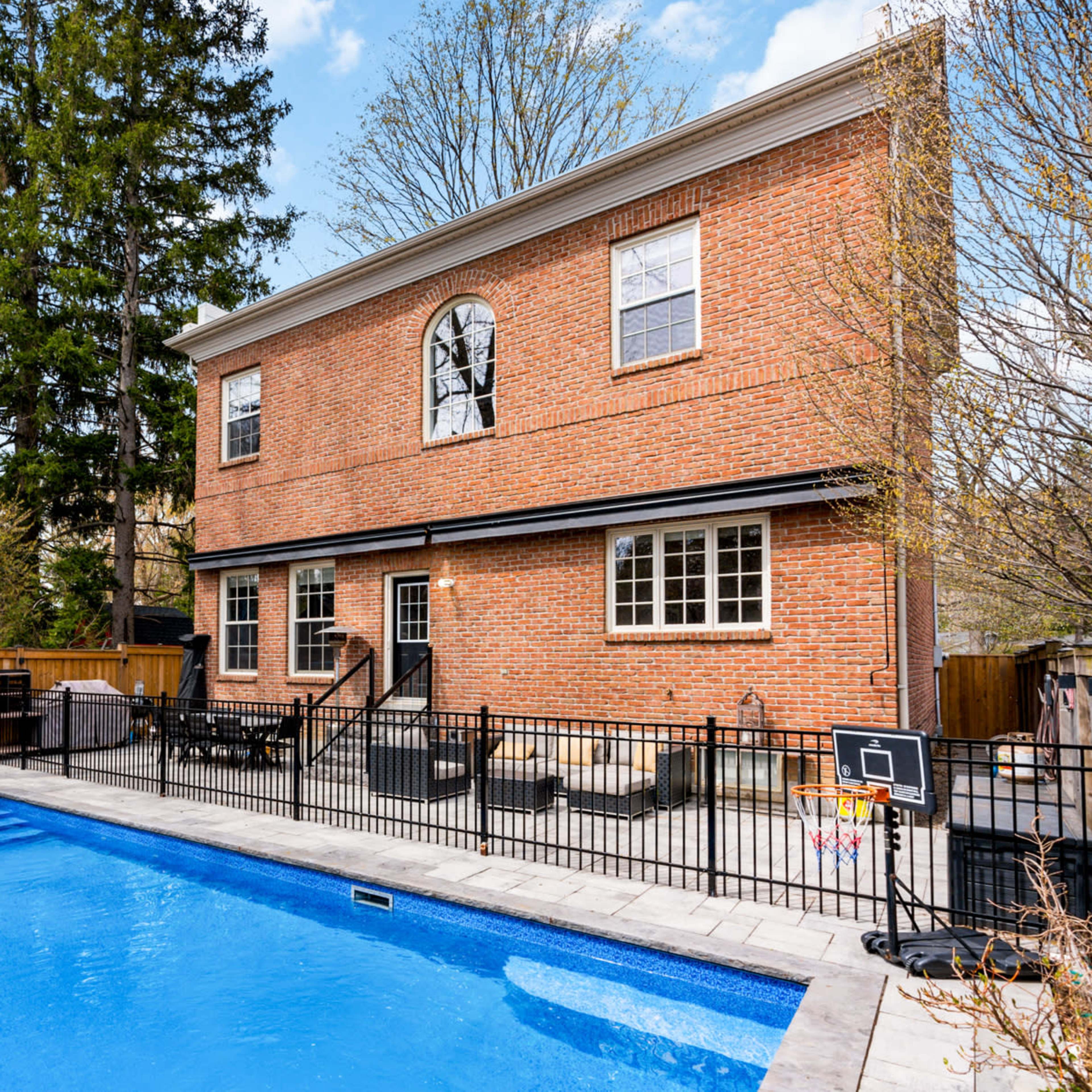 A two-story brick house with large windows overlooks a rectangular swimming pool surrounded by a patio and outdoor seating.