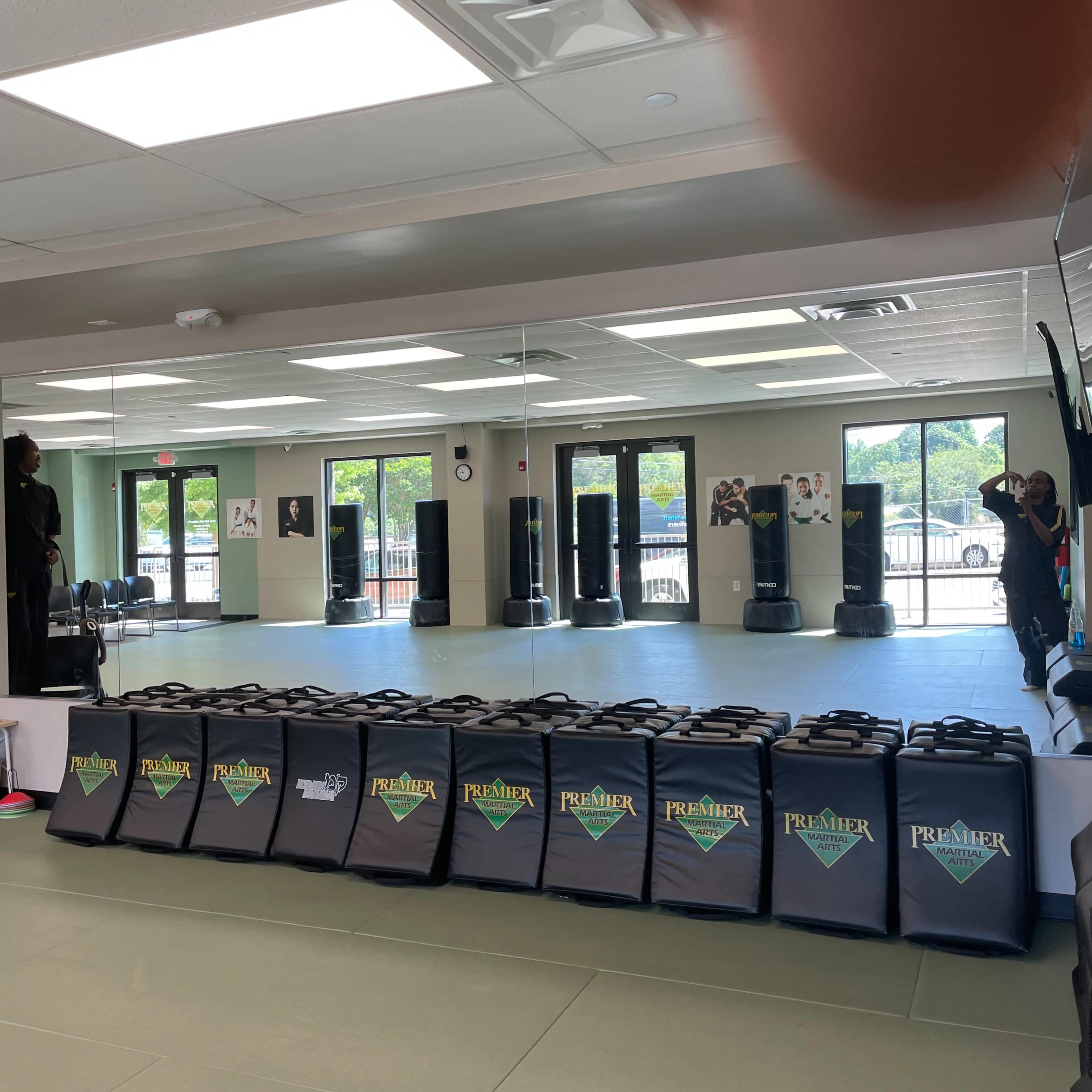 An empty martial arts studio with a mirrored wall, punching bags in the background, and training mats on the floor.
