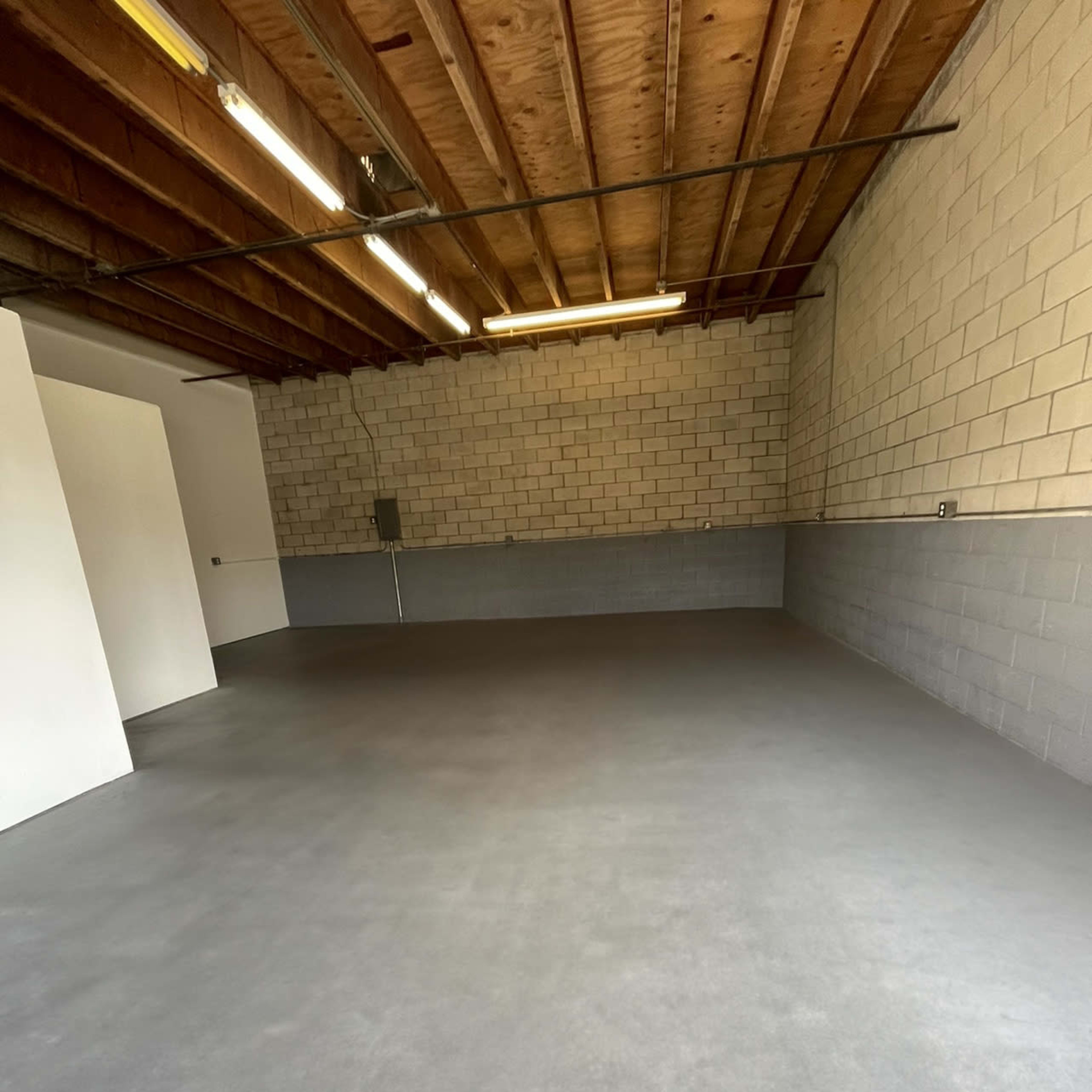 An empty garage or workshop space with concrete flooring, gray cinder block walls, and exposed wooden ceiling beams.