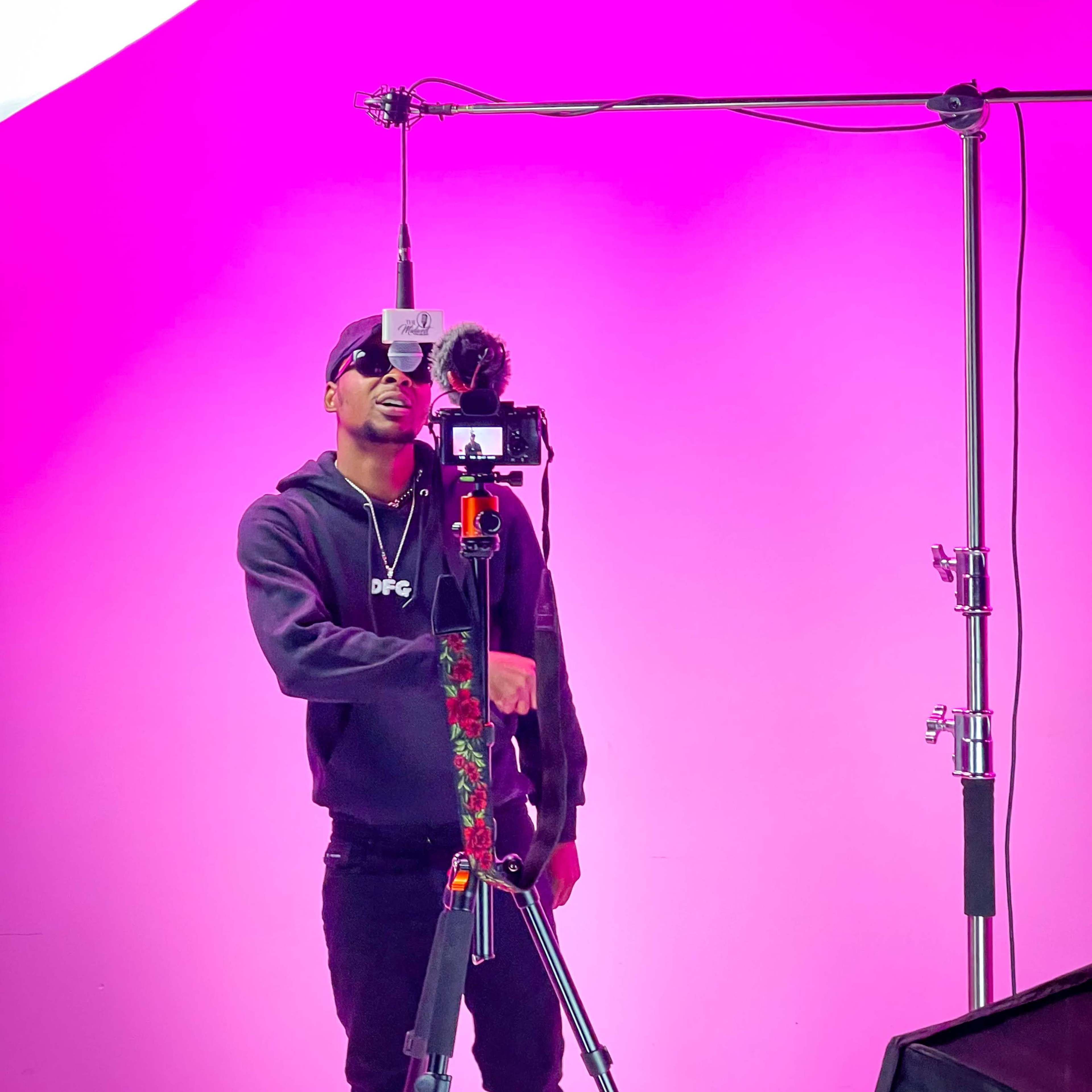 A person stands in front of a bright pink backdrop, adjusting a camera mounted on a tripod with a microphone attached.