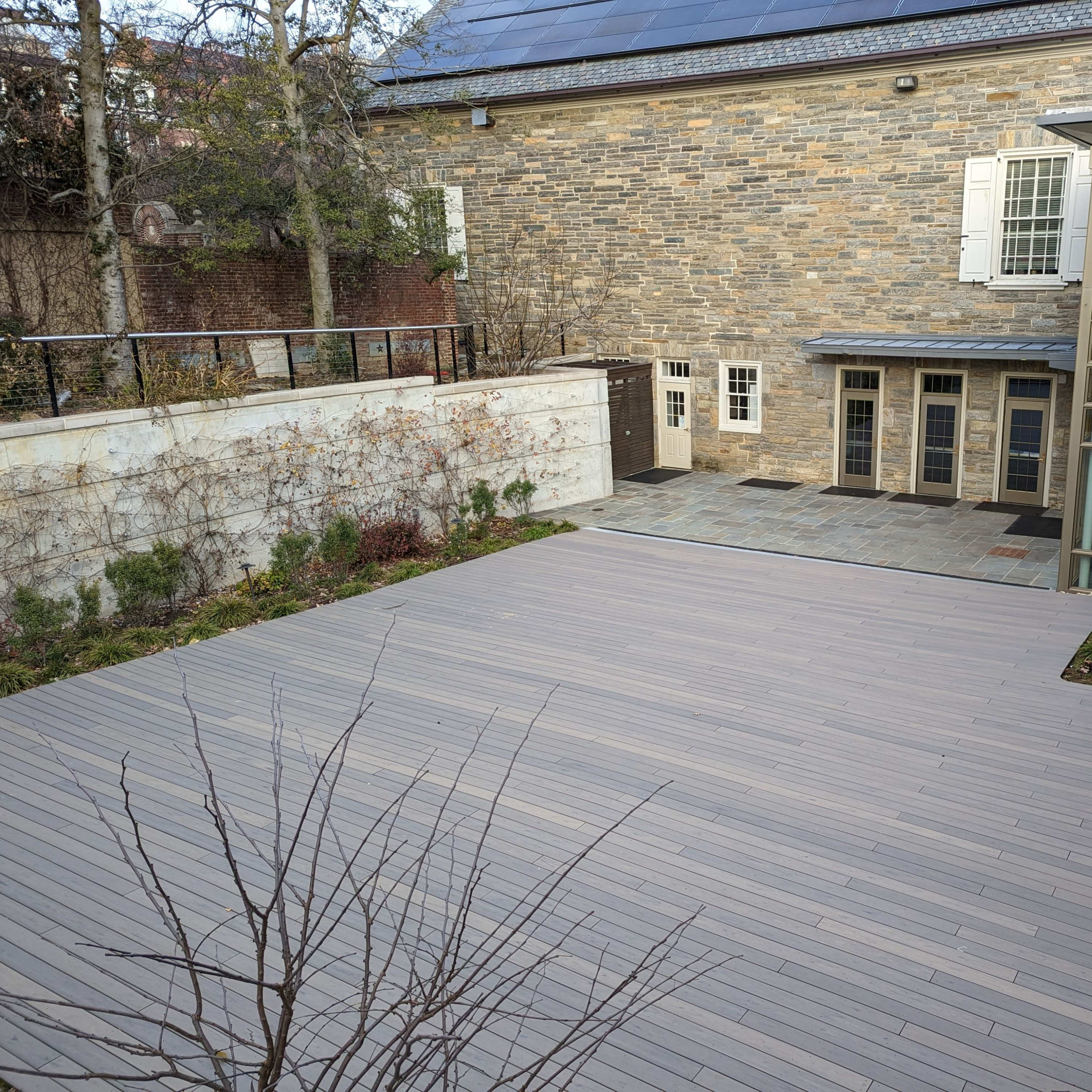 A modern outdoor terrace made of wooden decking adjacent to a stone building with large windows.