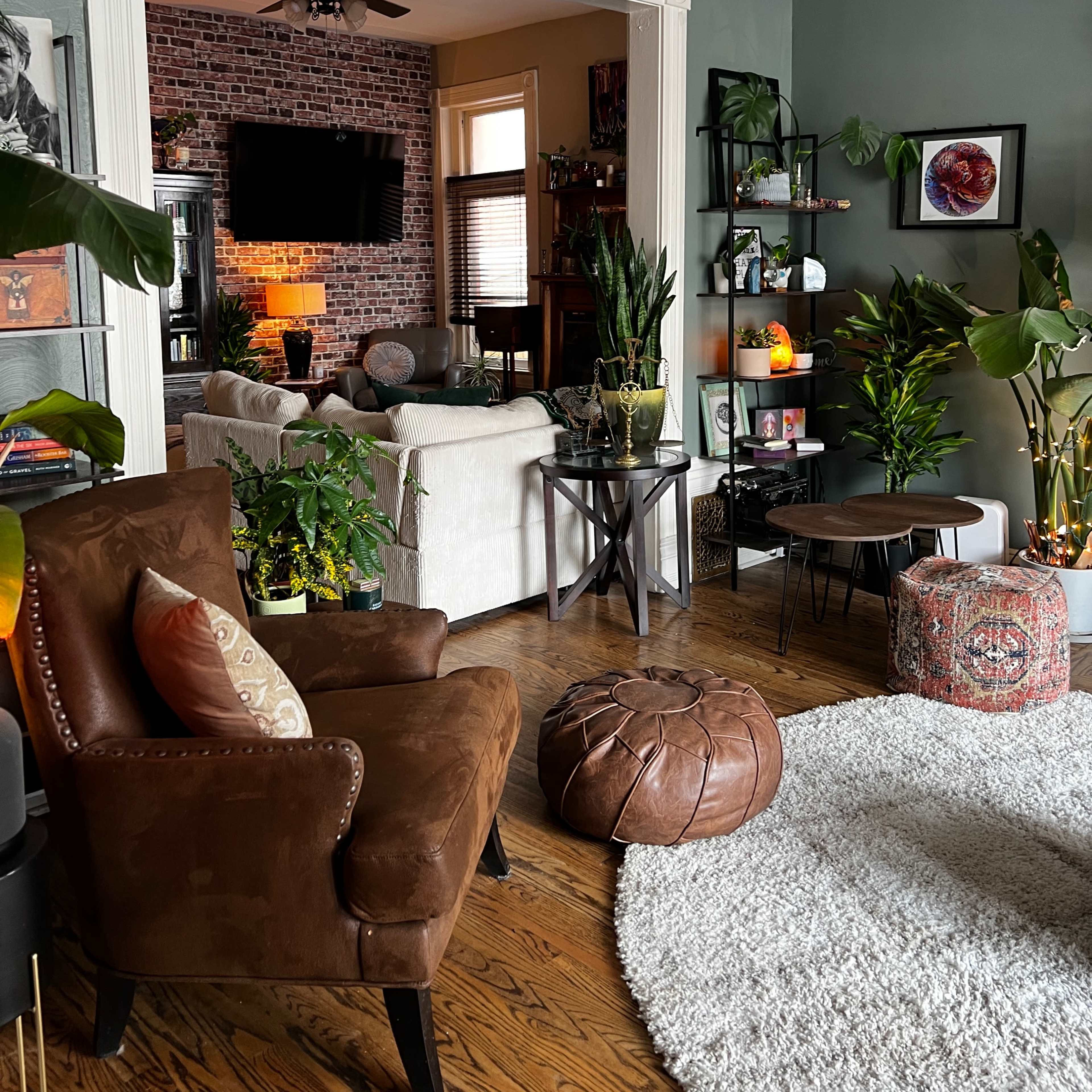 A cozy living room with a mix of plants, a brown armchair, a round leather pouf, and shelves displaying decorative items and books.
