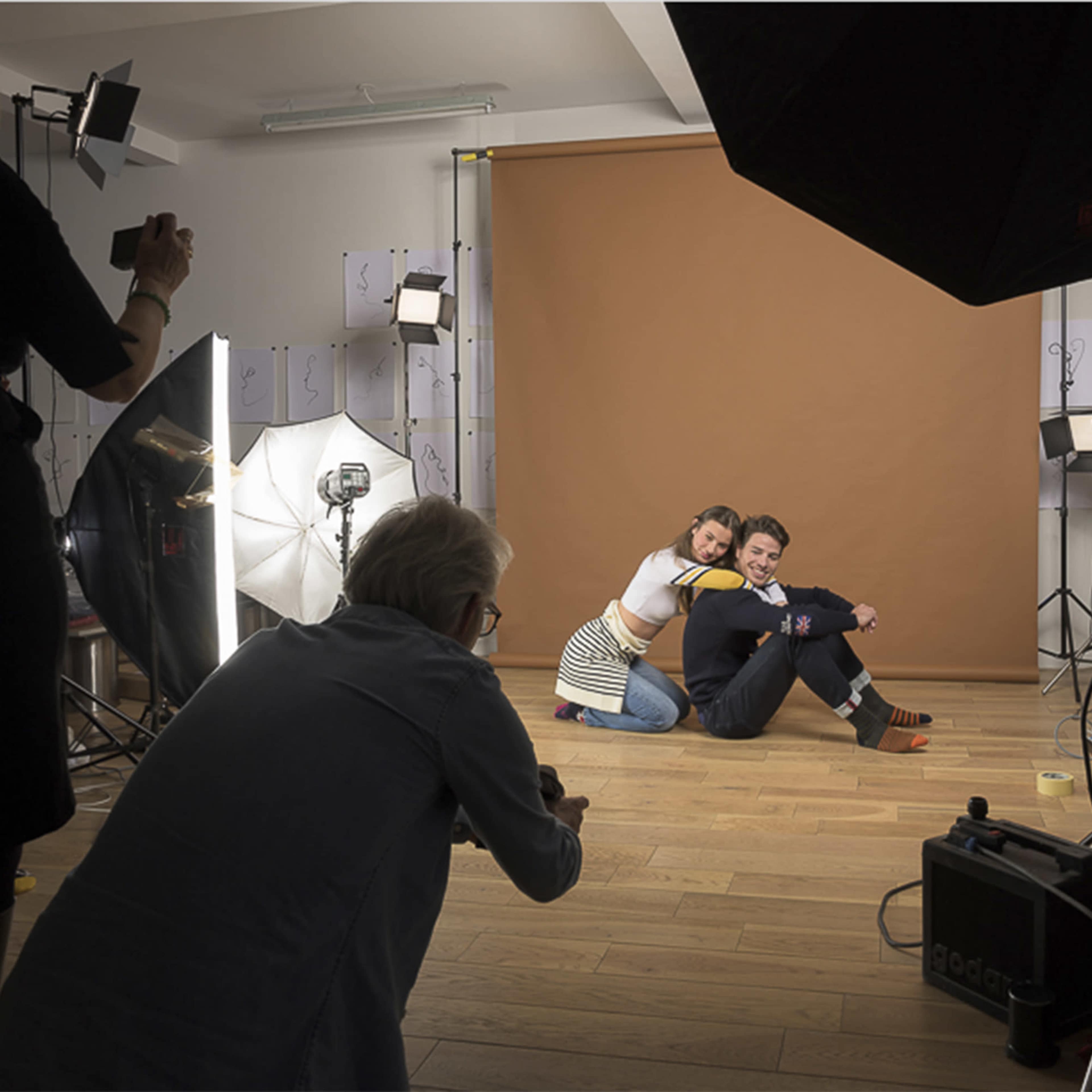 A photographer captures a couple posing together in a studio with a brown backdrop and various lighting equipment.