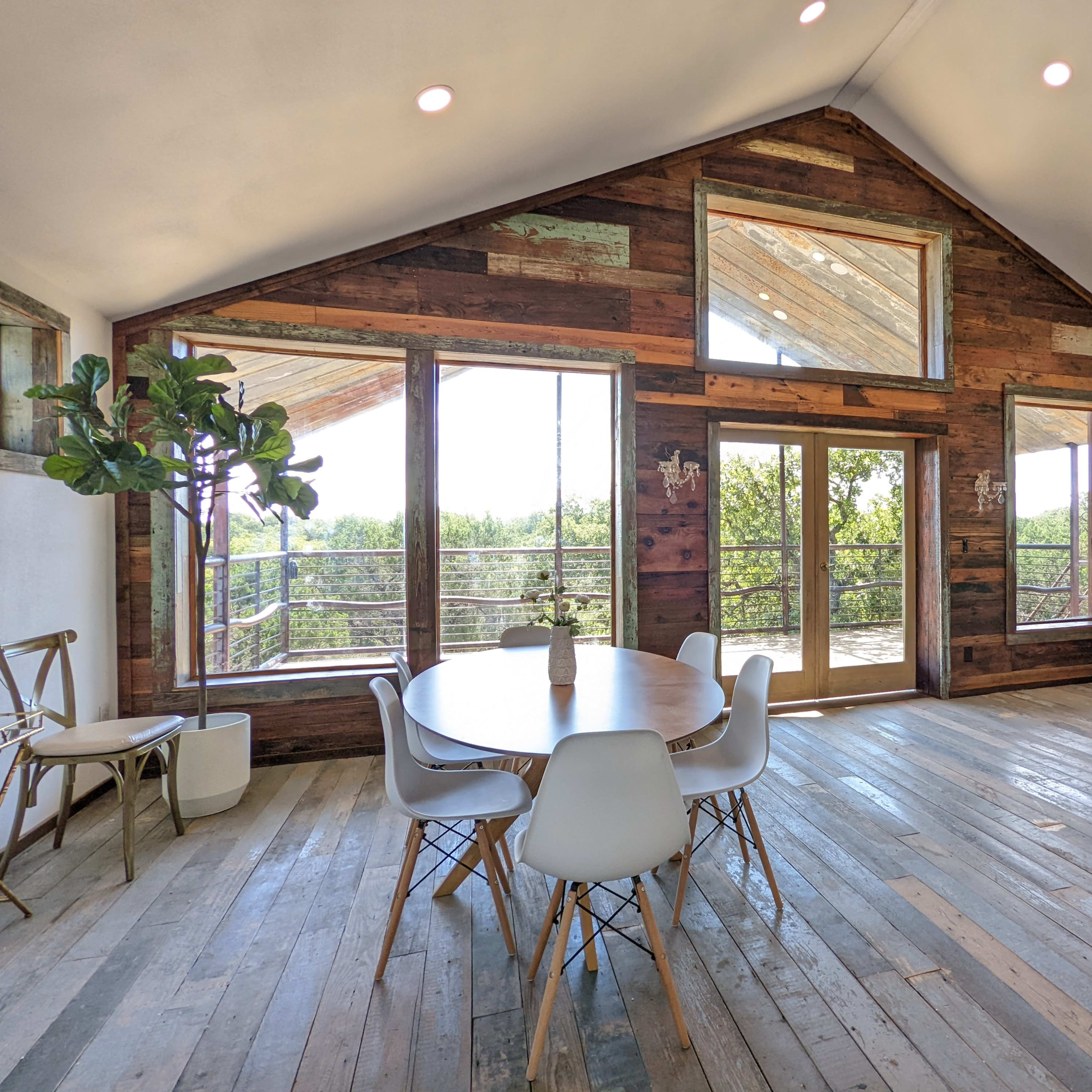 A spacious indoor area featuring a round dining table, modern chairs, large windows, and wooden walls, with a view of greenery outside.