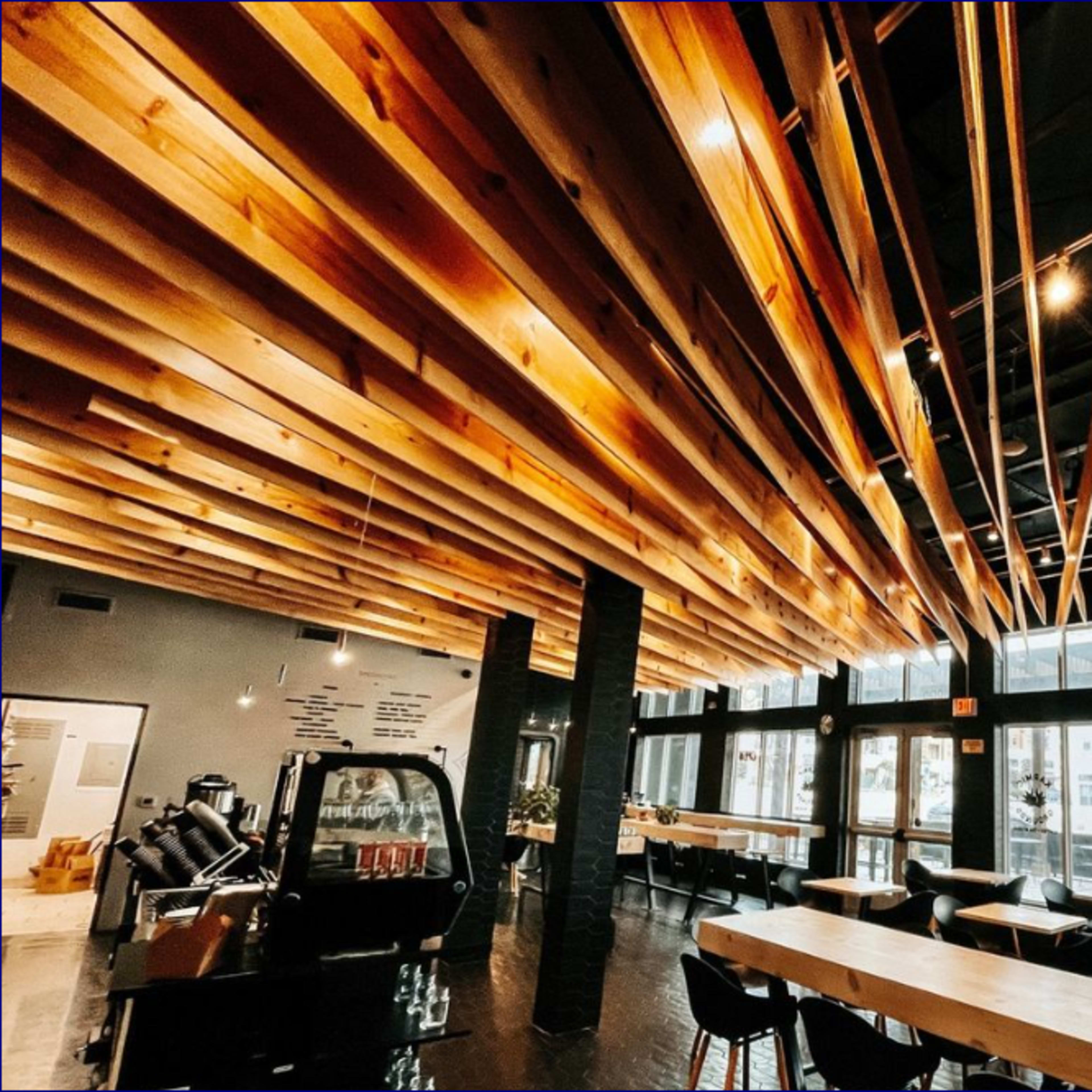 The interior of a modern cafe with a wooden slat ceiling, large windows, and a seating area with black chairs and wooden tables.