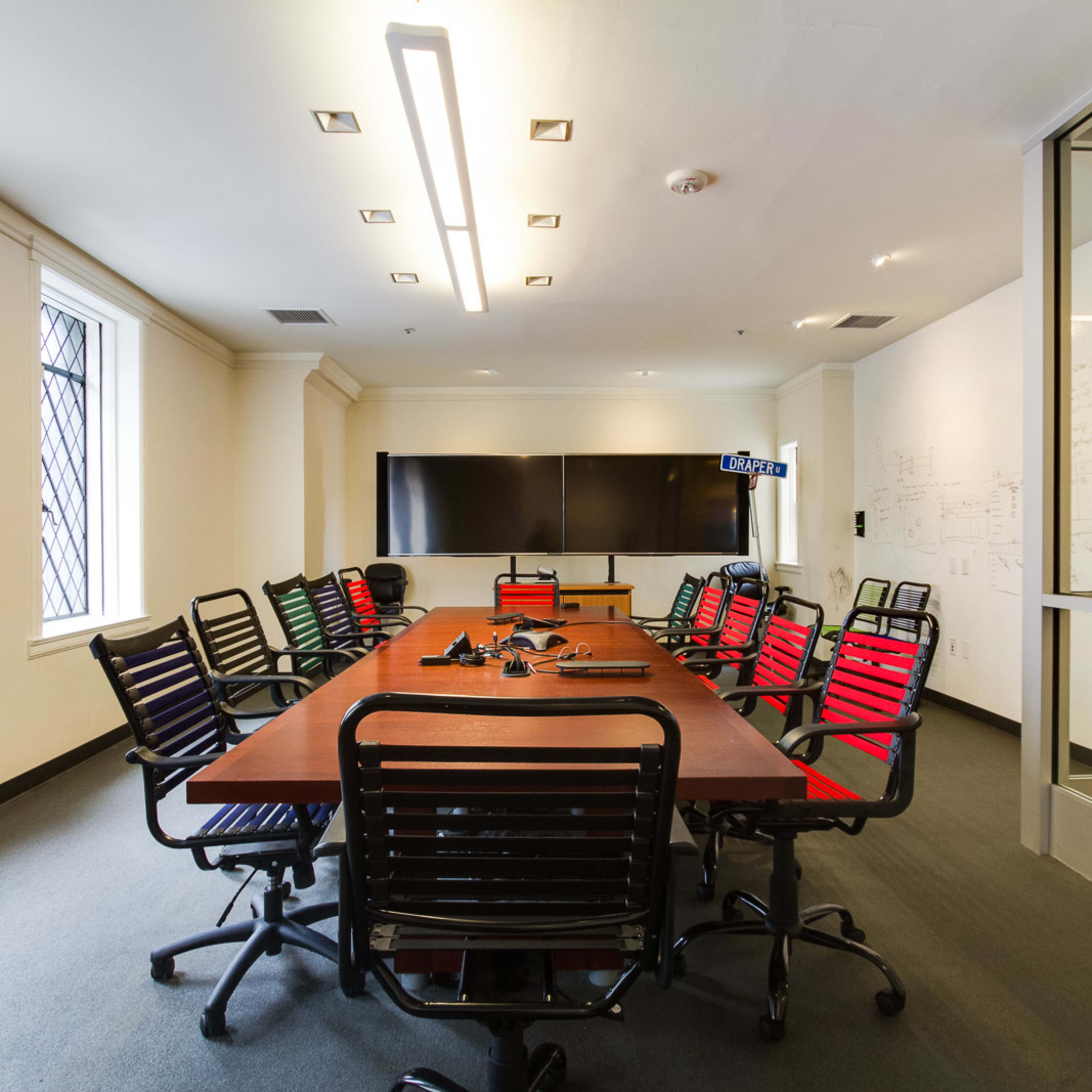 A conference room features a large wooden table surrounded by colorful office chairs and equipped with a large screen and whiteboard.