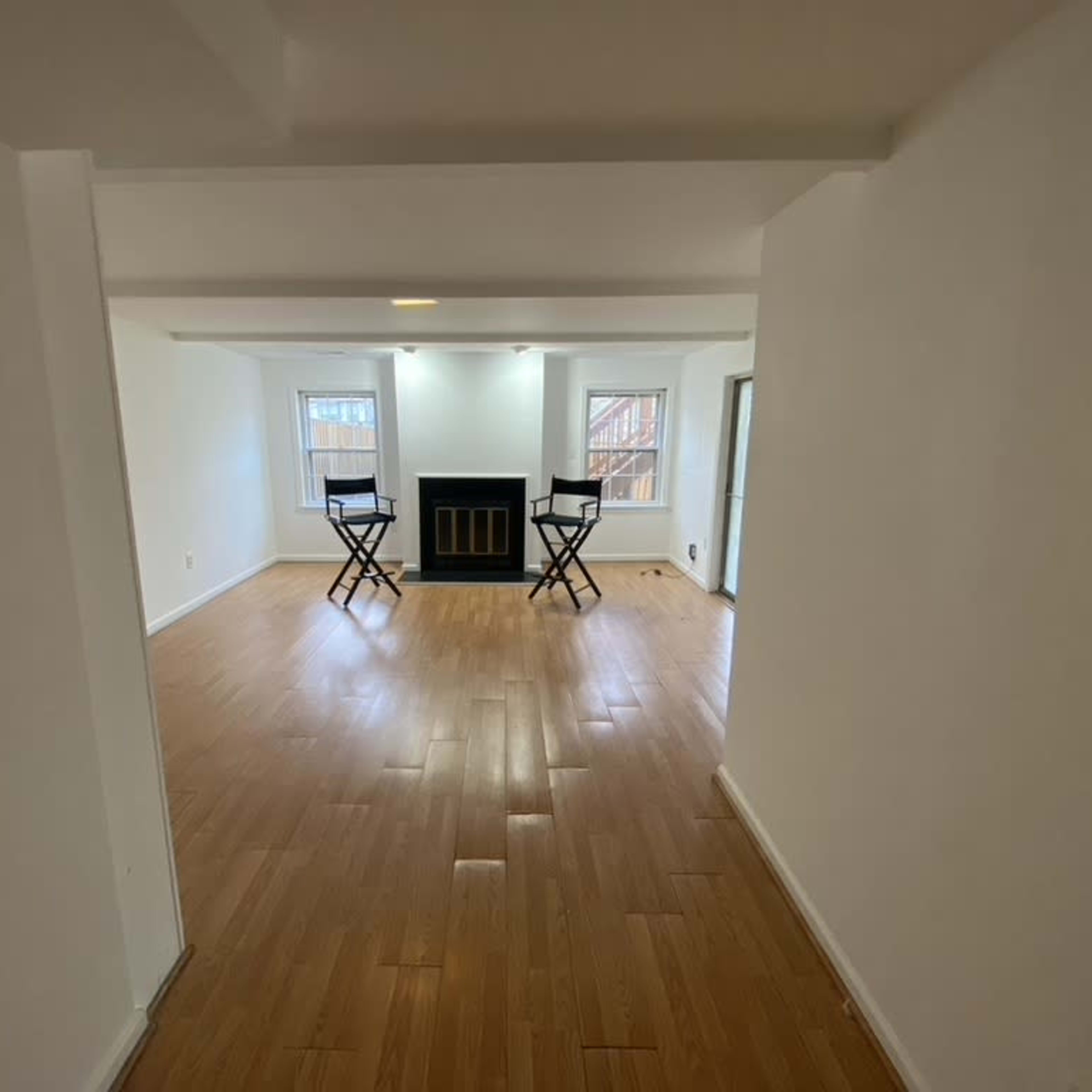A spacious, empty room with wooden flooring, two black stools facing a fireplace, and large windows letting in natural light.