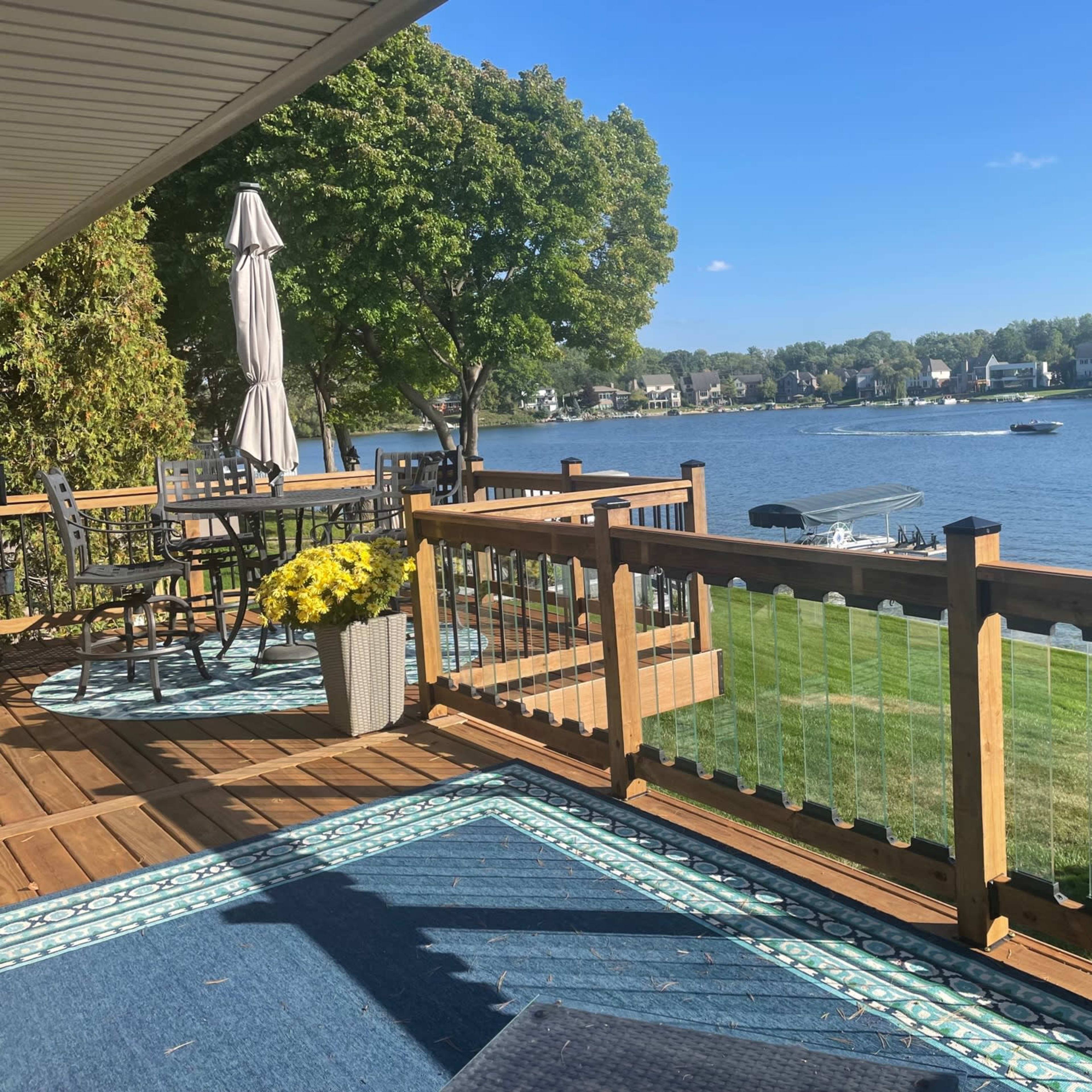 A wooden deck overlooks a lake, featuring a patio table and chairs, an umbrella, and a boat passing by on the water.