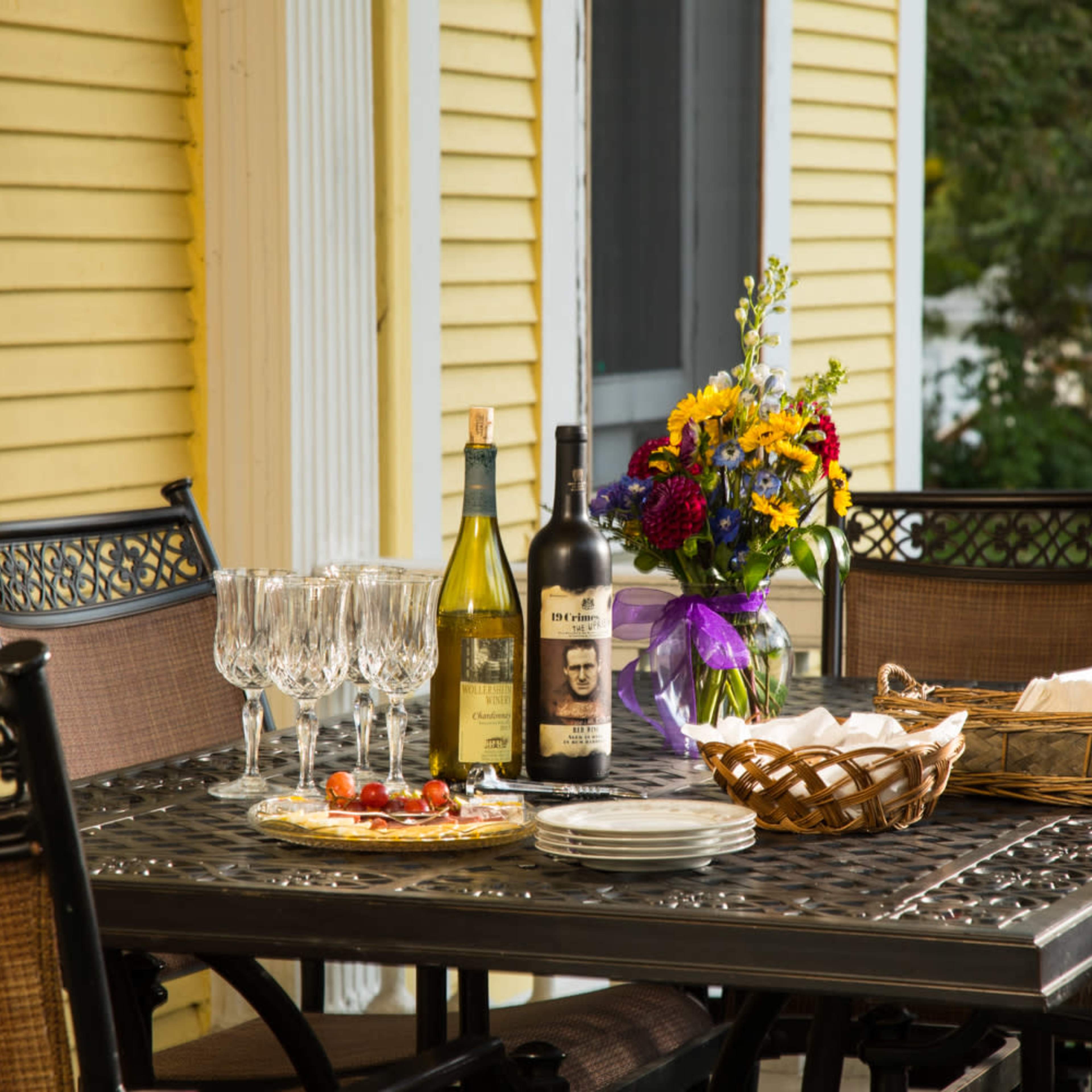 A table set for outdoor dining features wine bottles, glasses, a basket, plates, and a floral centerpiece.