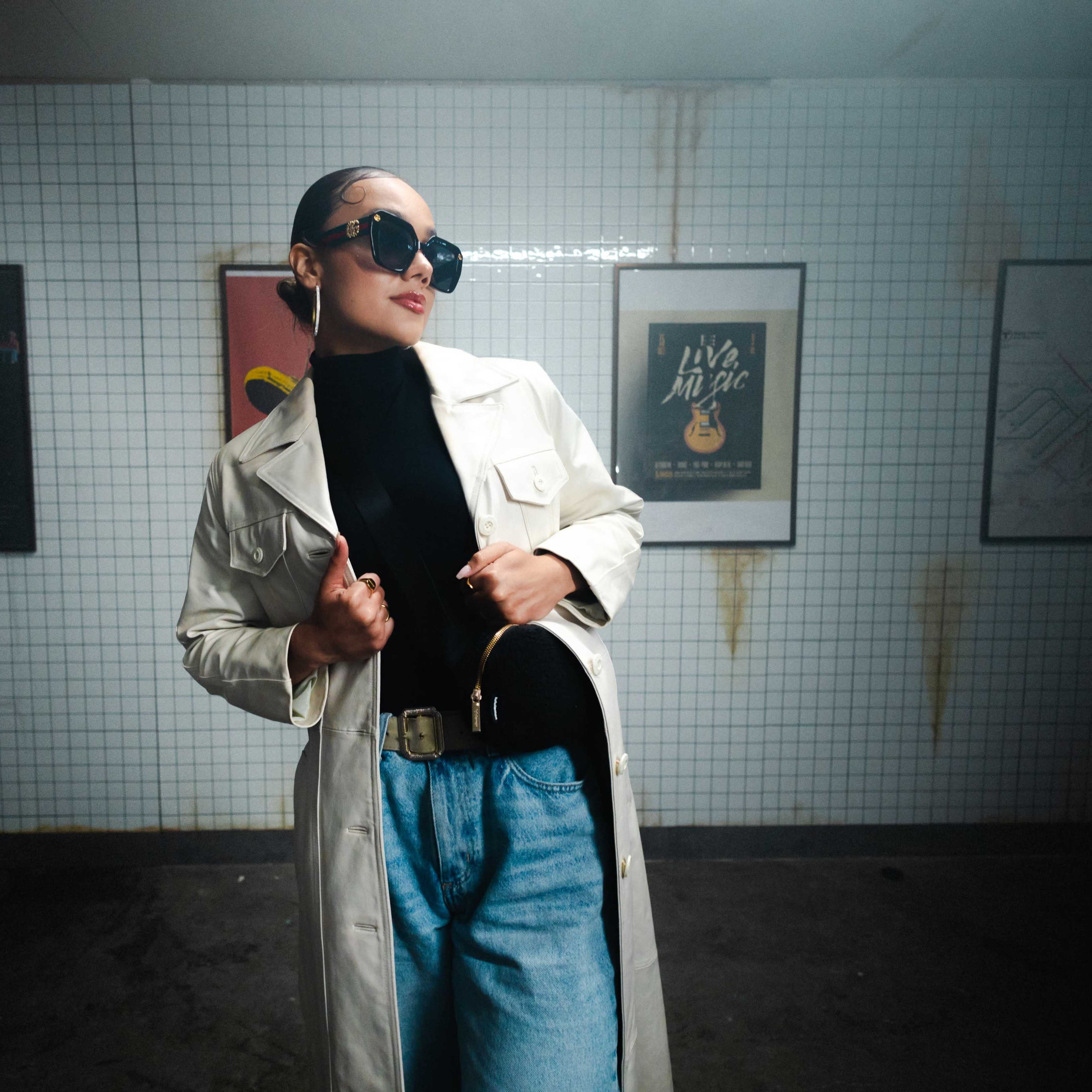 A person wearing a white trench coat and sunglasses poses against a tiled wall with framed posters.