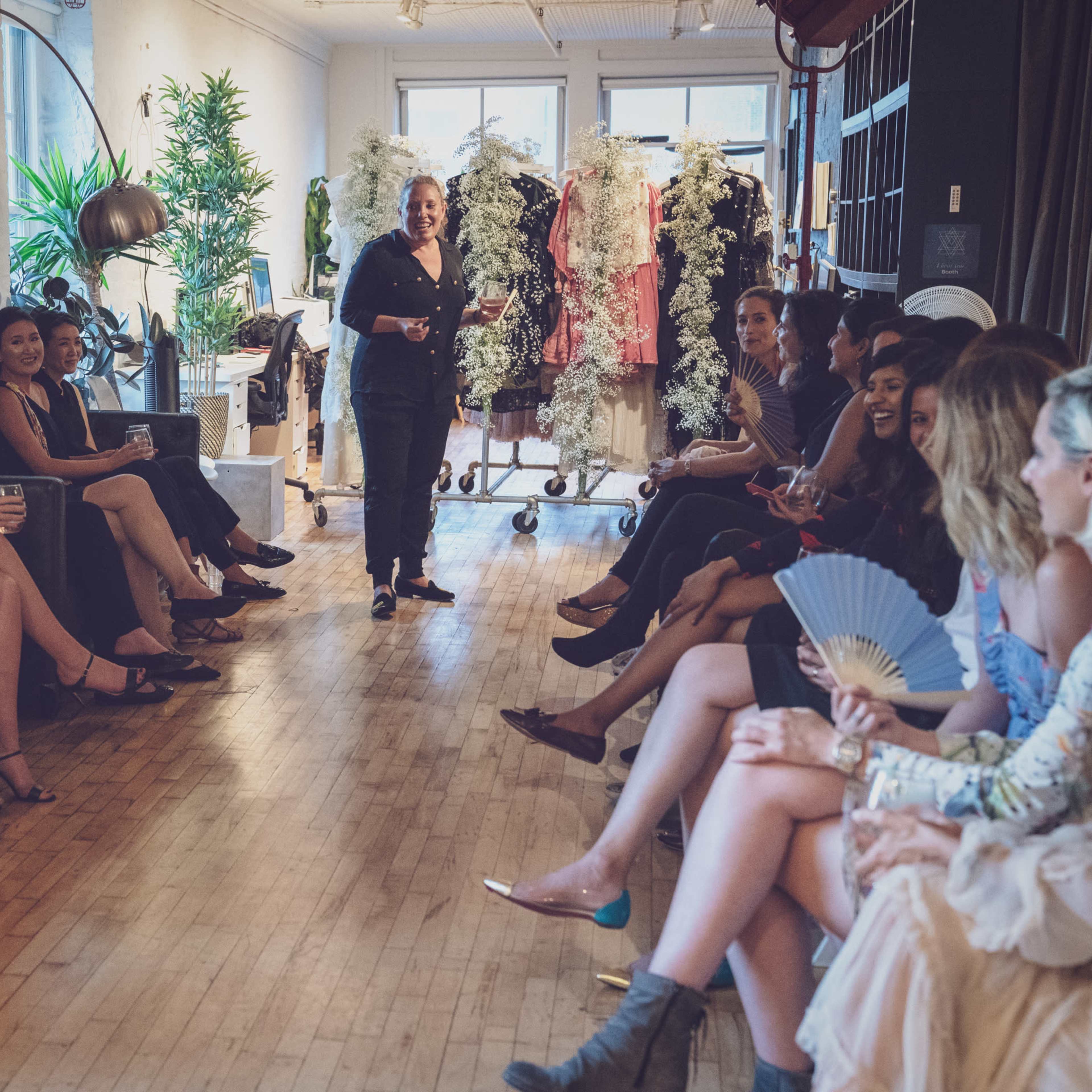 A group of women sits on one side of a stylish room while a speaker addresses them in front of racks of clothing.