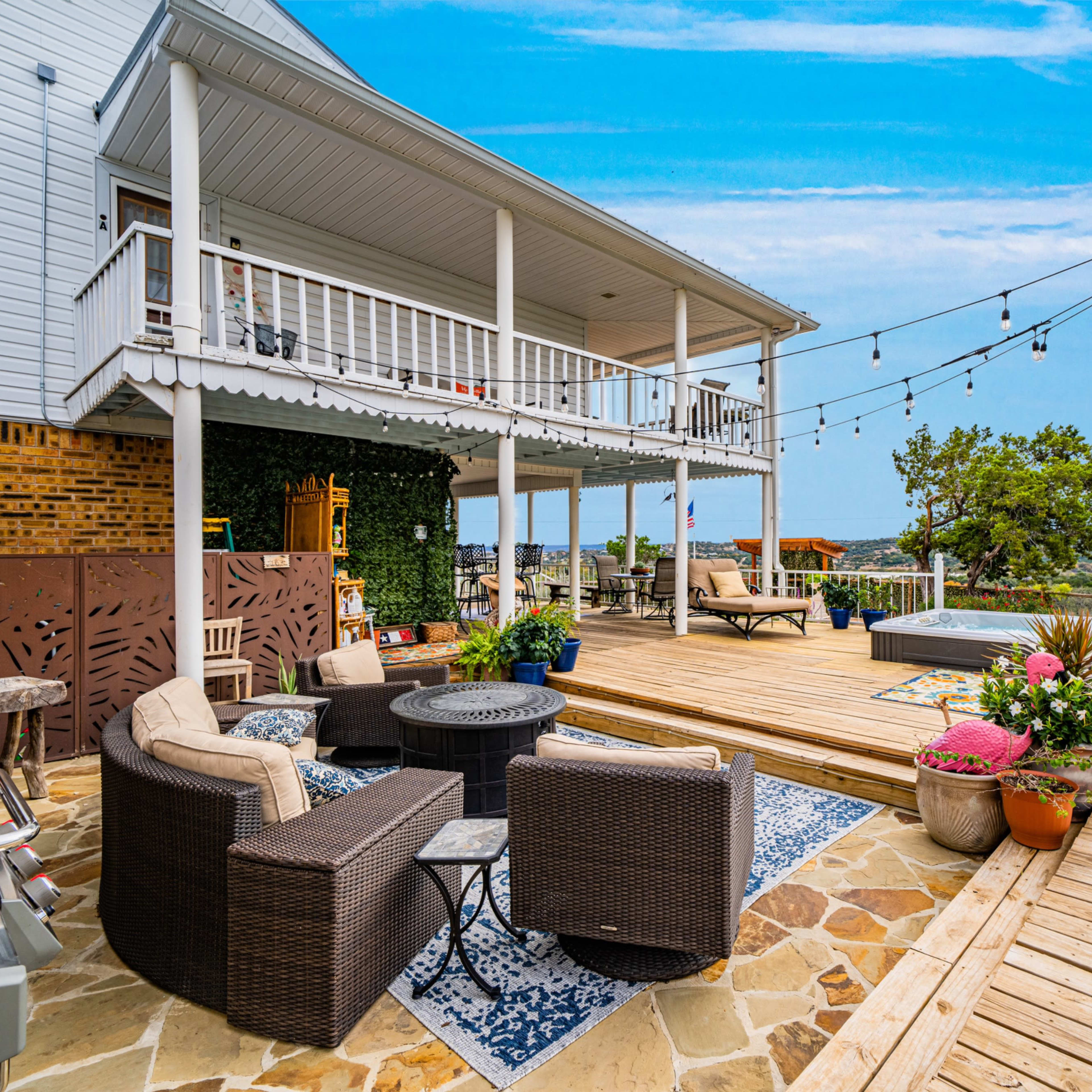A wooden deck with outdoor furniture, including a seating area and a grill, surrounded by potted plants and string lights, with a view of a landscape in the background.