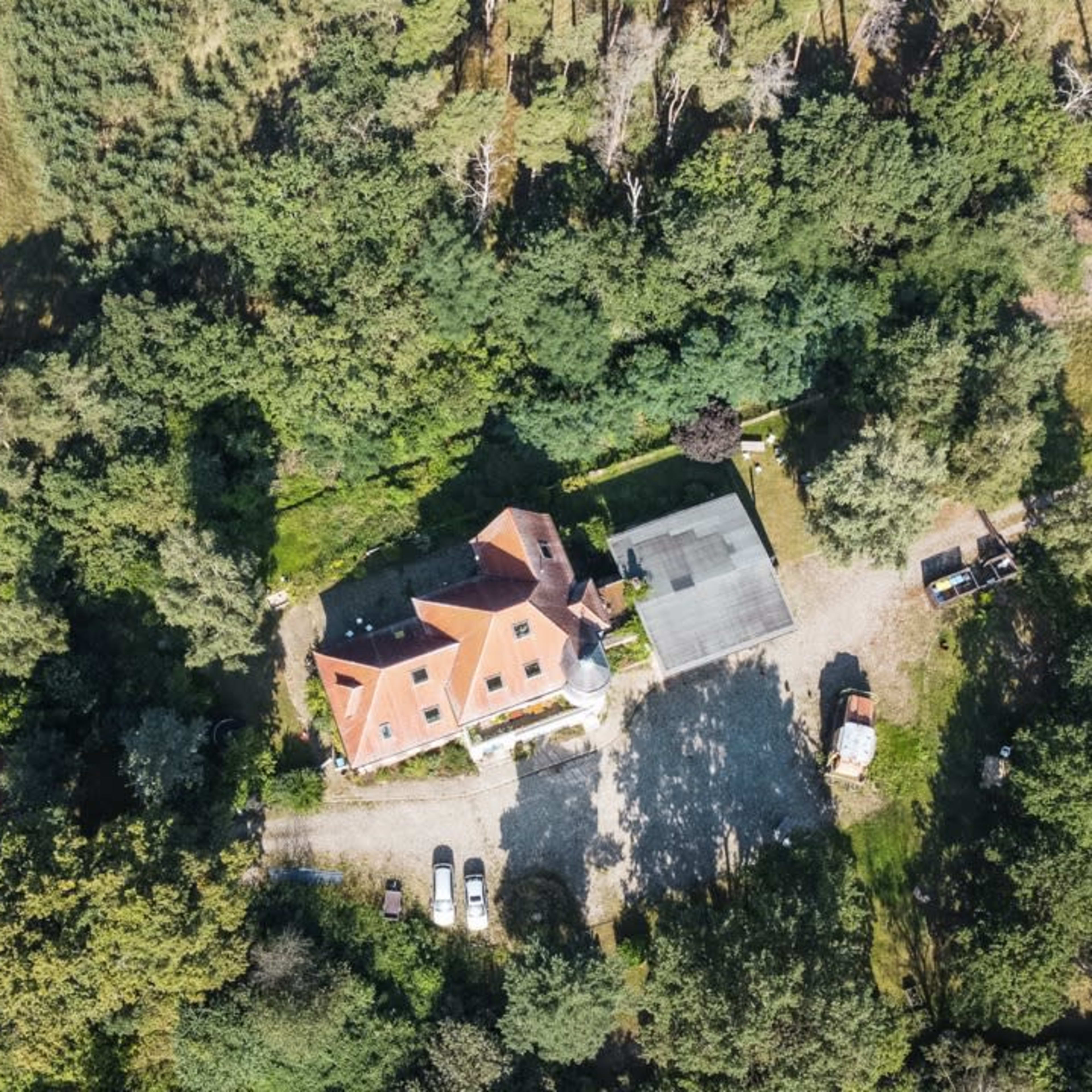 An aerial view shows a house surrounded by dense greenery, with a gravel driveway leading to the property and additional parked vehicles nearby.