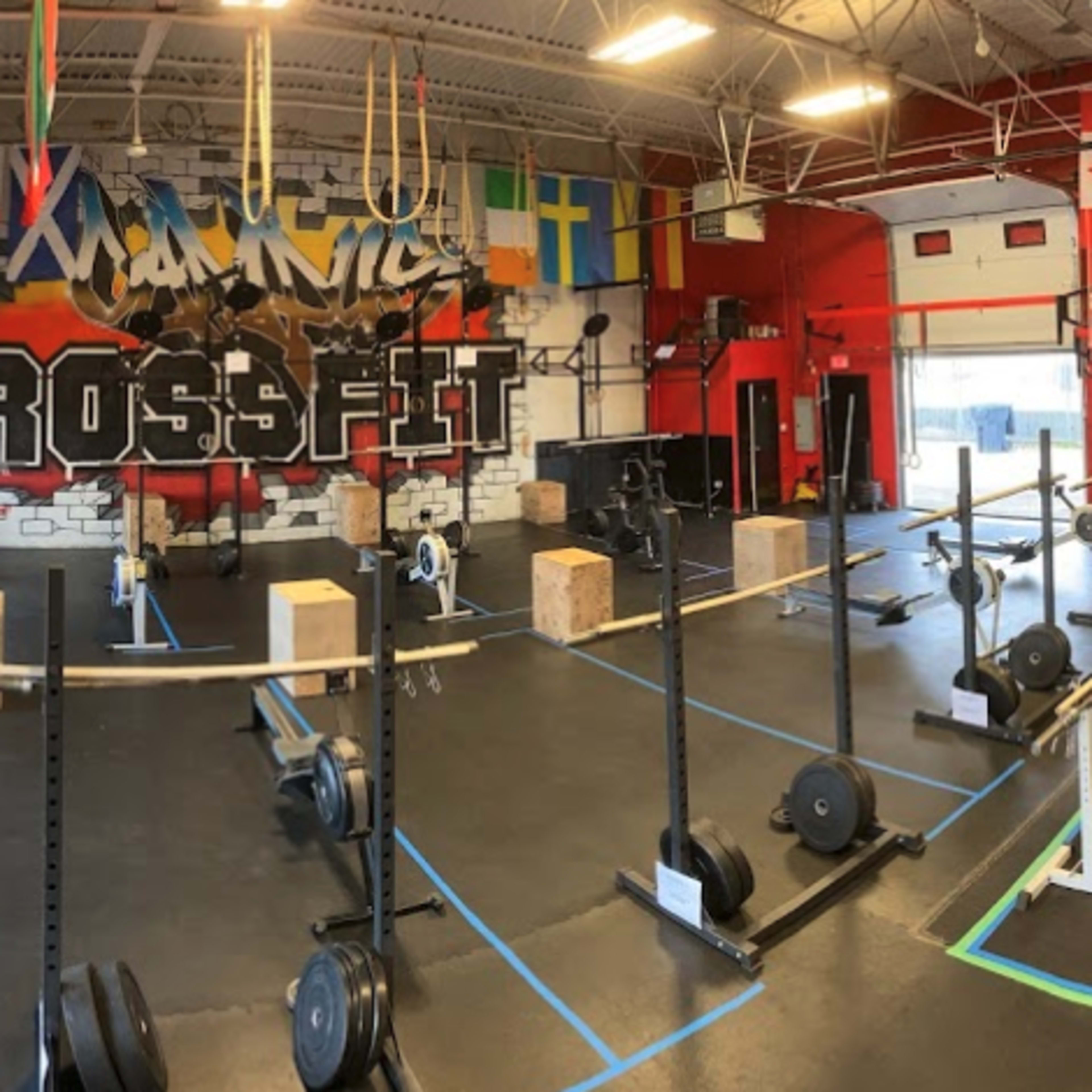 A CrossFit gym with various workout equipment arranged on a black rubber floor, illuminated by overhead lights and decorated with flags and artwork on the walls.