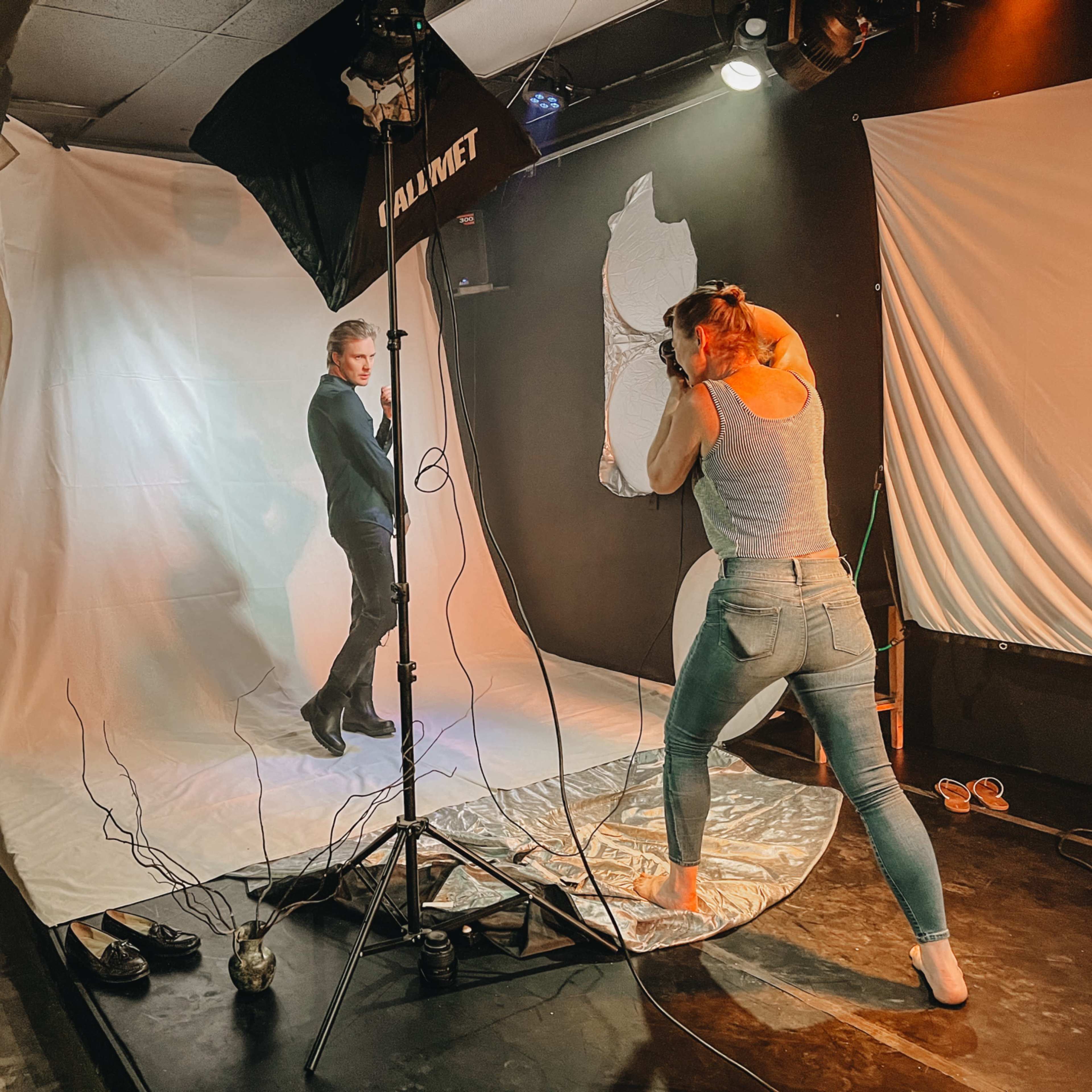 A photographer captures a model posing against a plain backdrop in a studio setting, with lighting equipment and props arranged around them.