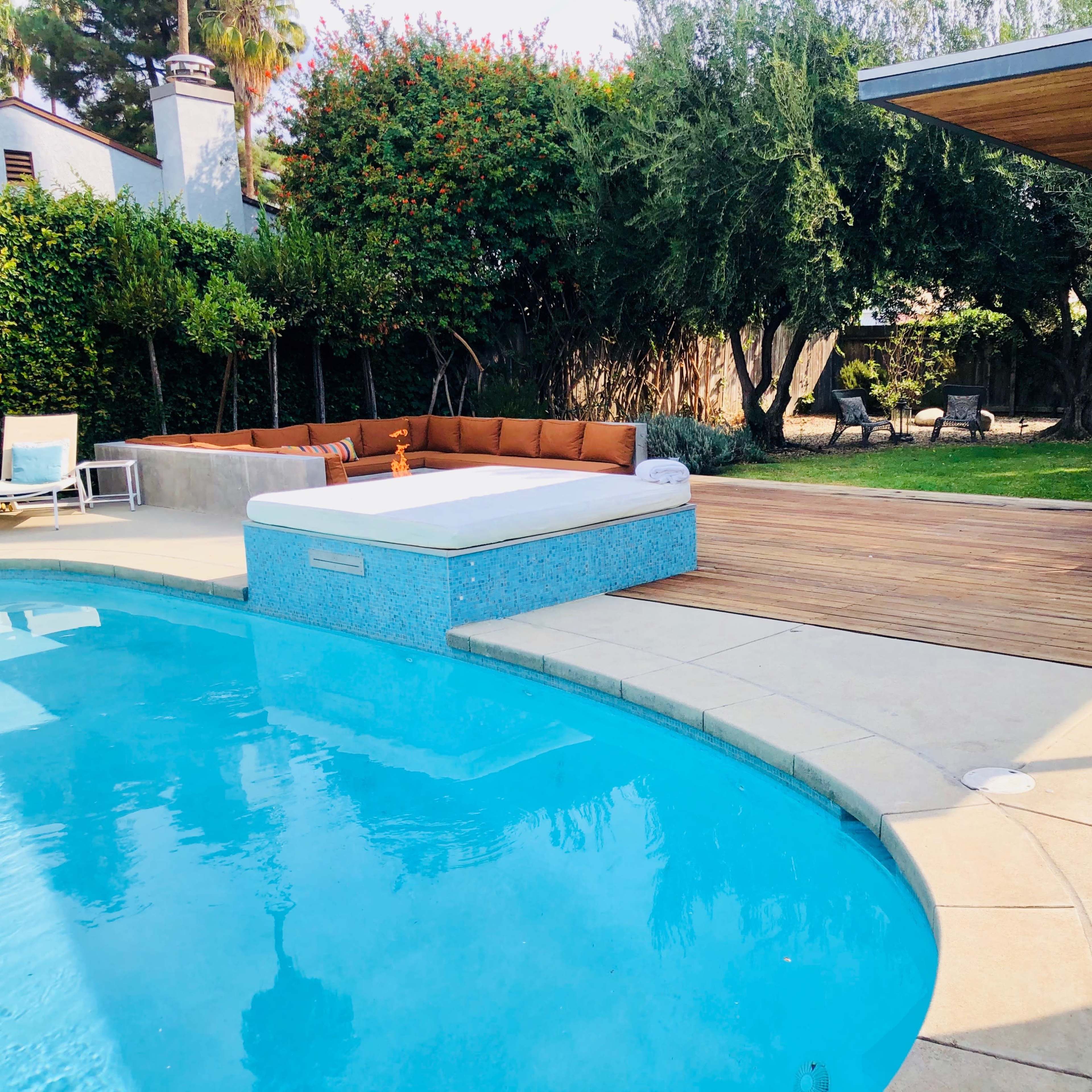 A swimming pool surrounded by a wooden deck and outdoor seating area with a covered lounge space in a backyard setting.