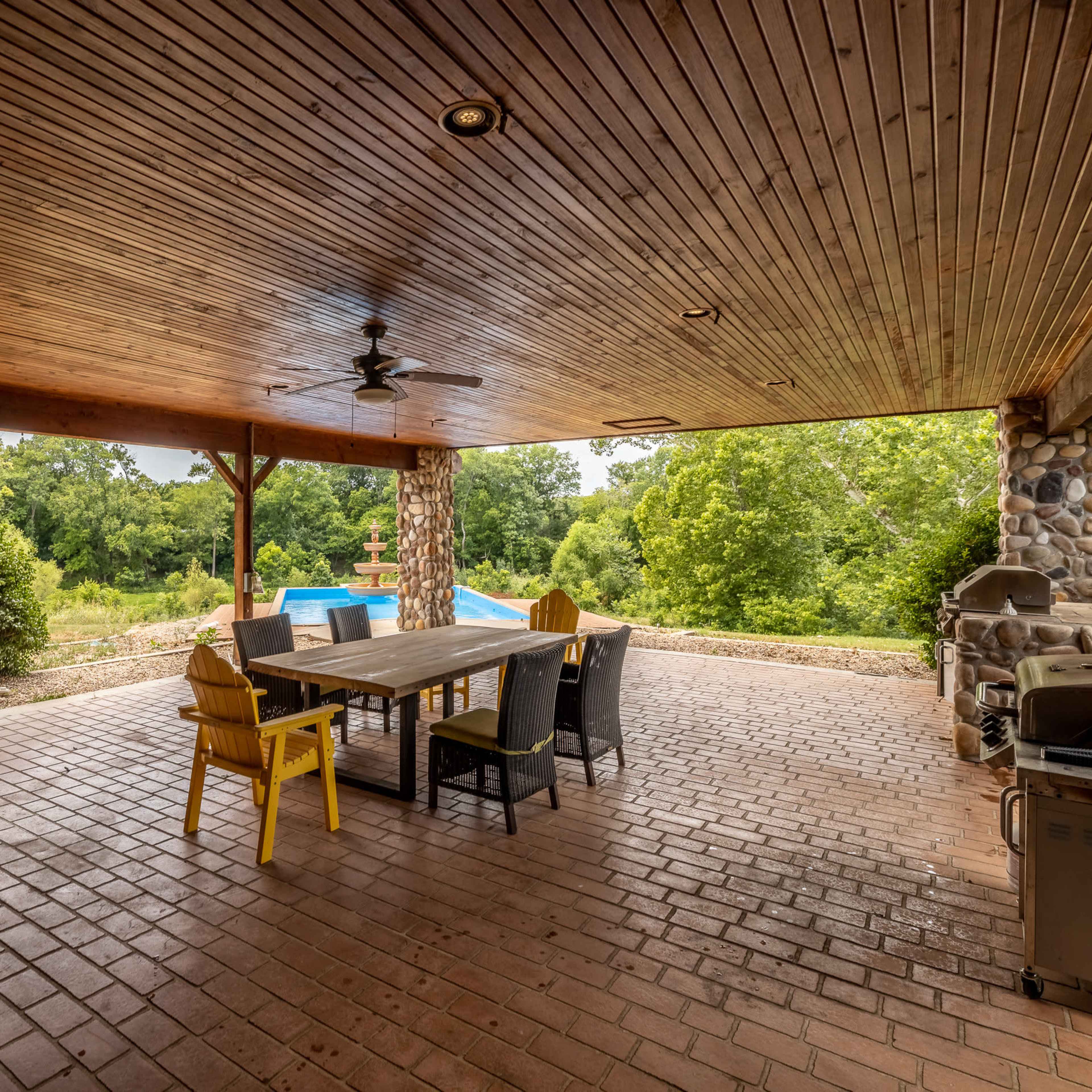 A covered outdoor patio area featuring a wooden dining table surrounded by chairs, a barbecue grill, and a view of greenery and a pool in the background.