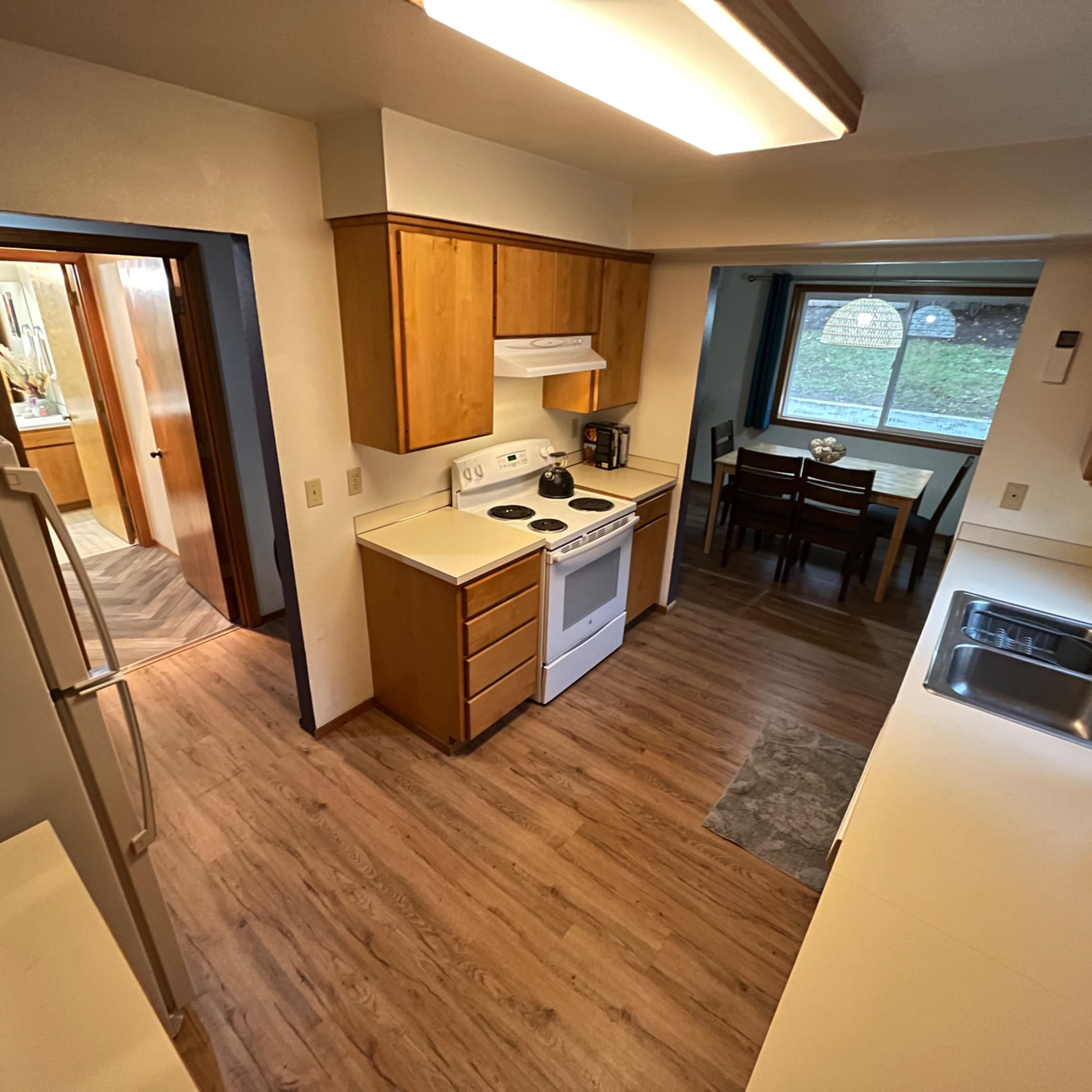 A kitchen with wooden cabinets, a stove, and a dining area visible through a window, leading to a bathroom door in the background.