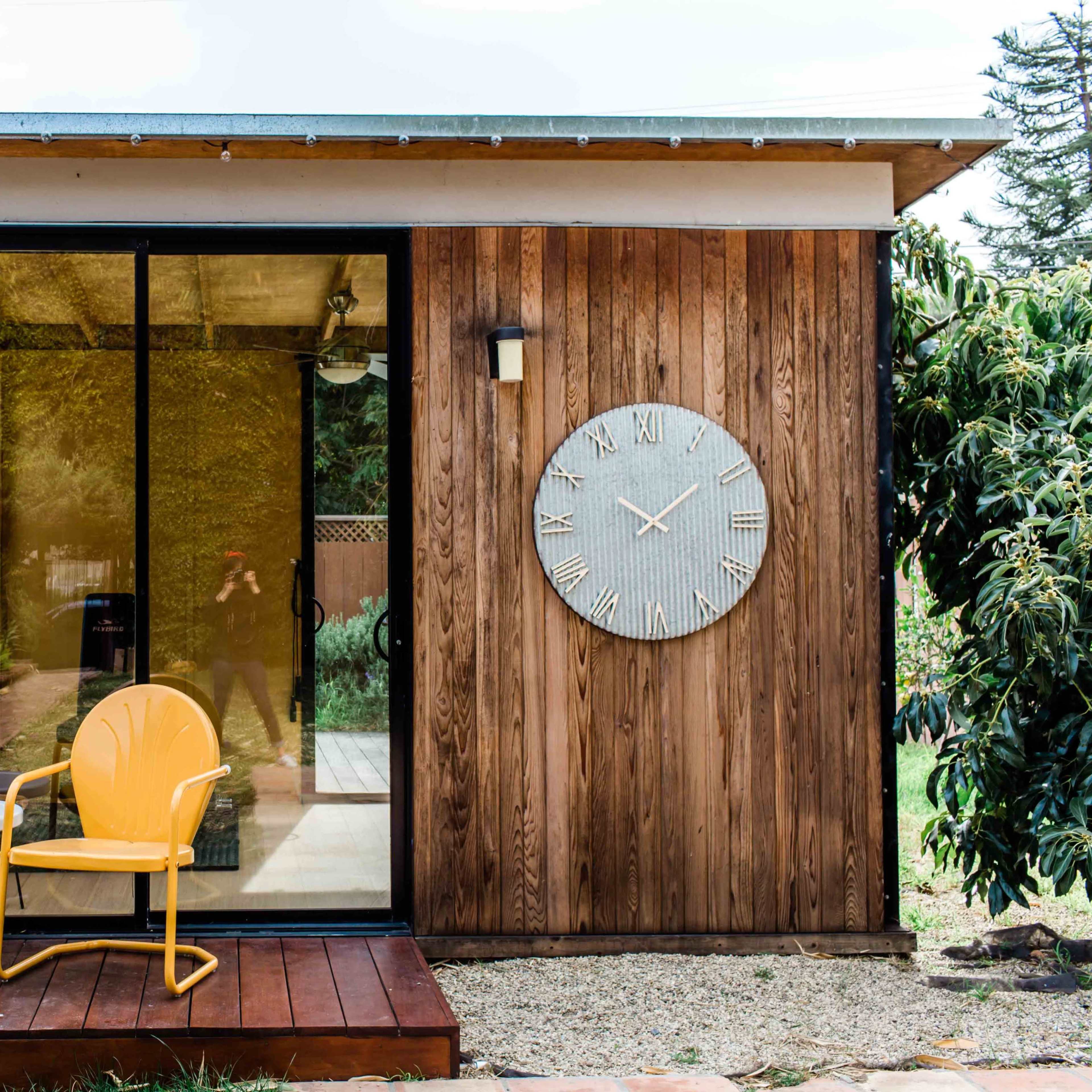 A small wooden structure with a large clock on the wall, featuring two yellow chairs and a round table on a wooden deck in front.