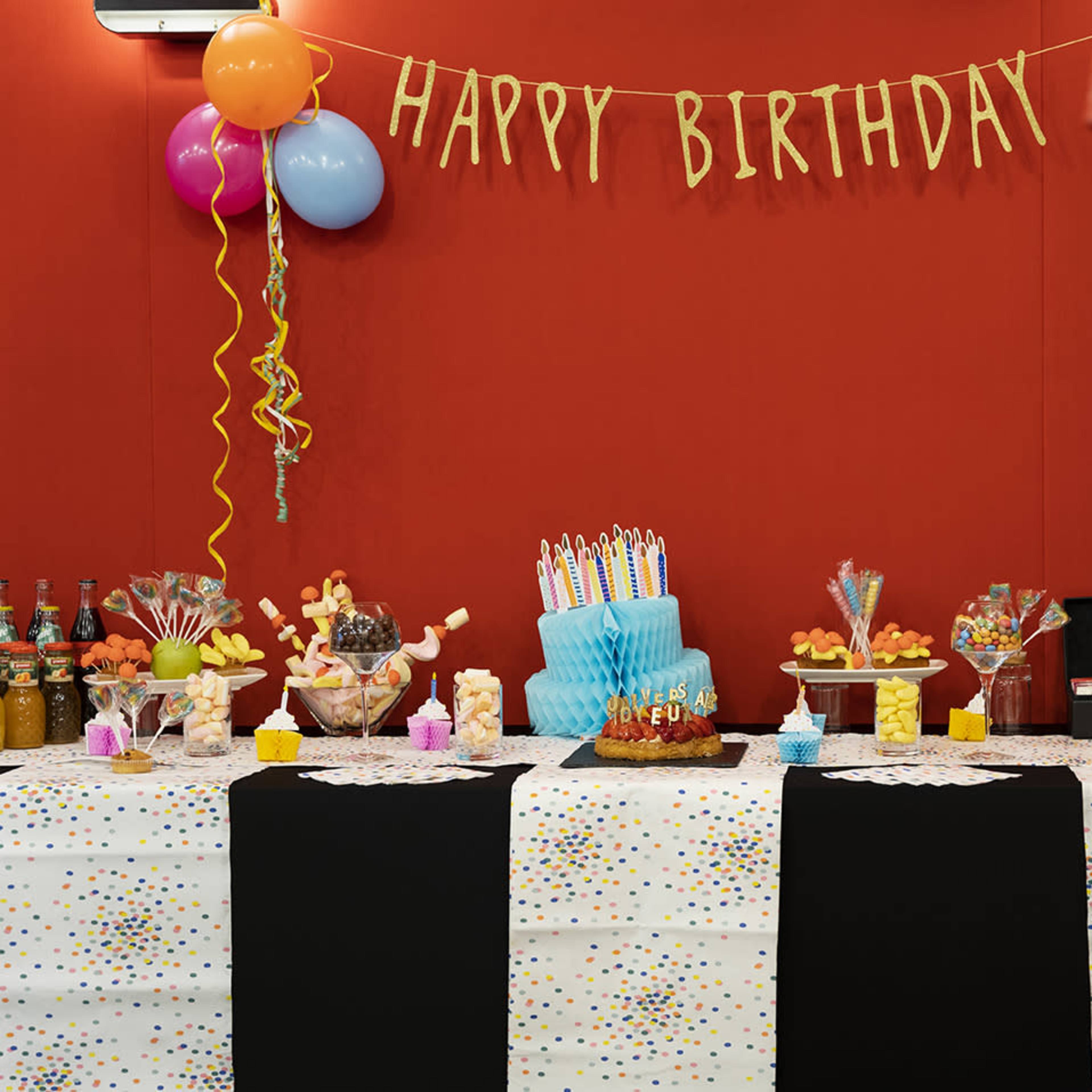 A birthday celebration setup features a table adorned with colorful decorations, a blue cake with candles, and a popcorn machine in the foreground.