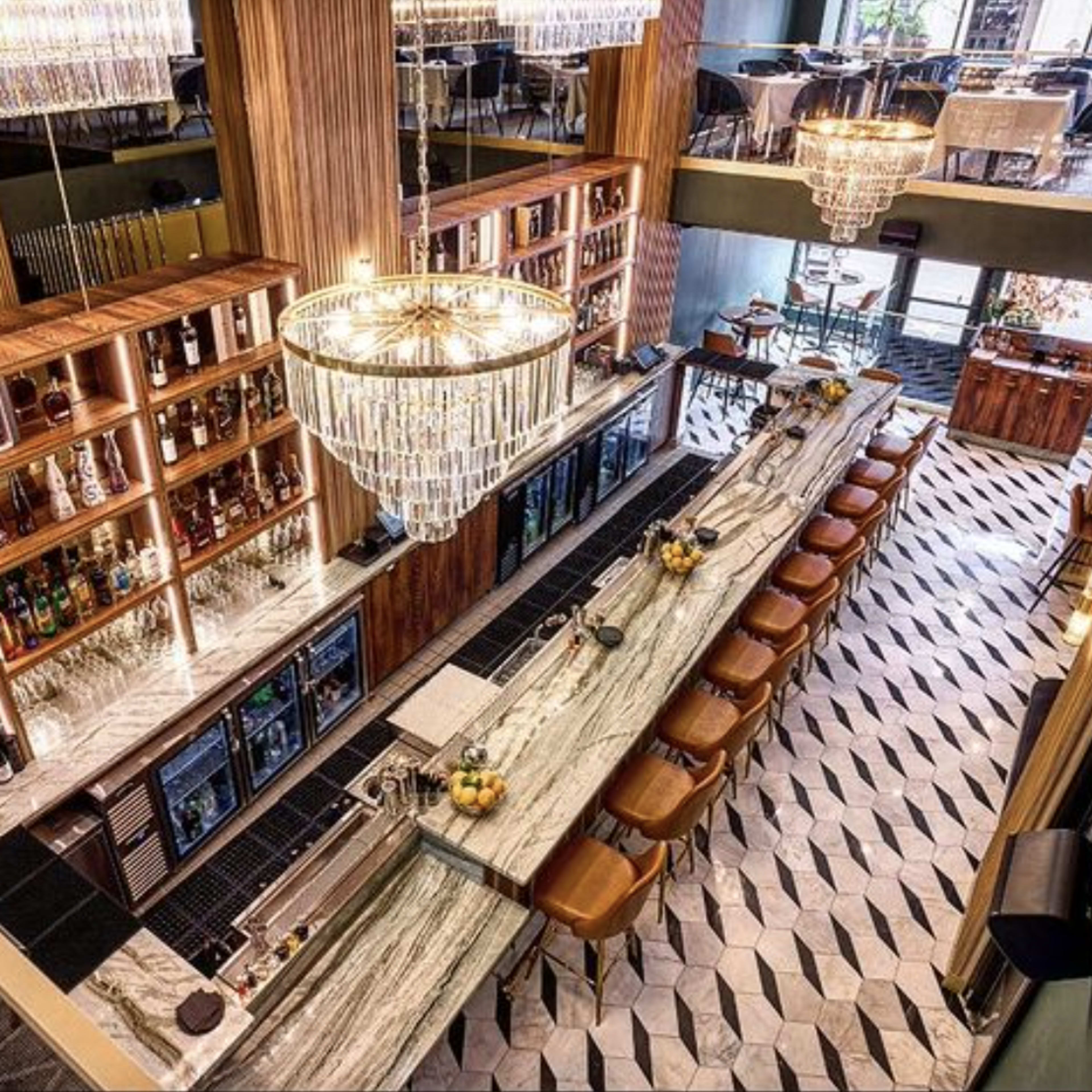 A spacious bar with a long marble countertop, surrounded by seating and illuminated by large chandeliers.