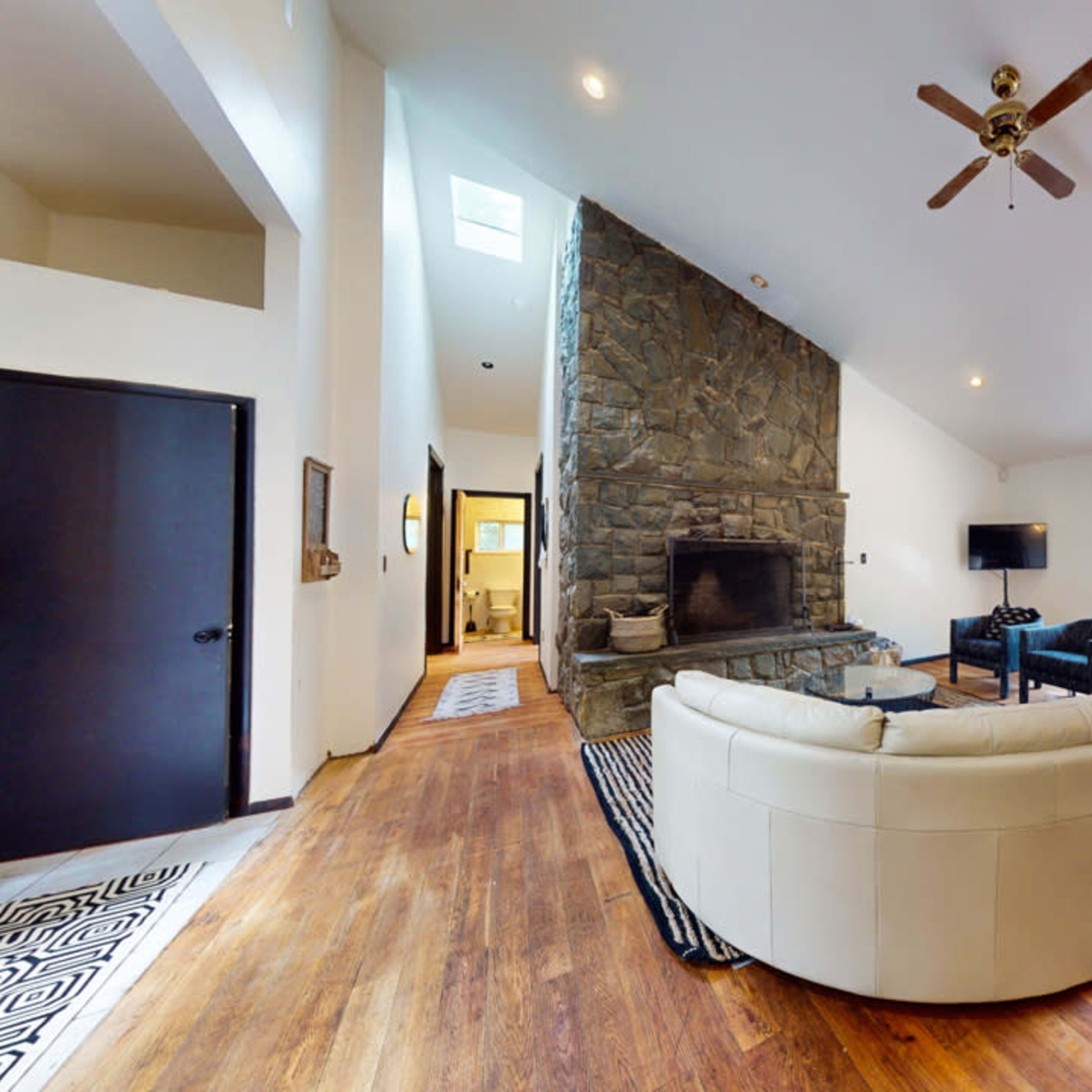 A spacious living room featuring a large stone fireplace, a curved sectional sofa, and windows that provide a view of surrounding greenery.