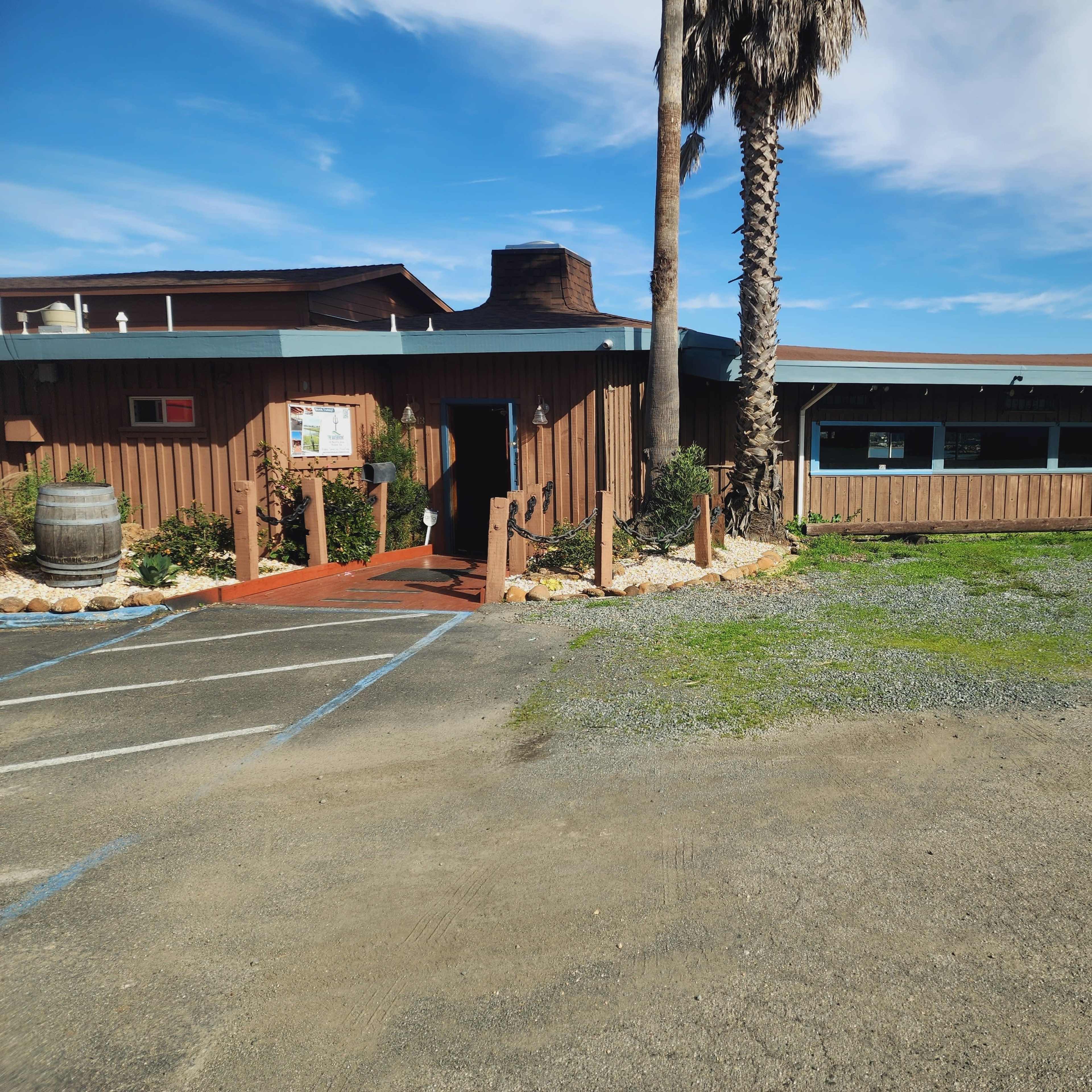 A wooden building with a patio area and palm tree stands next to a gravel parking lot under a clear sky.
