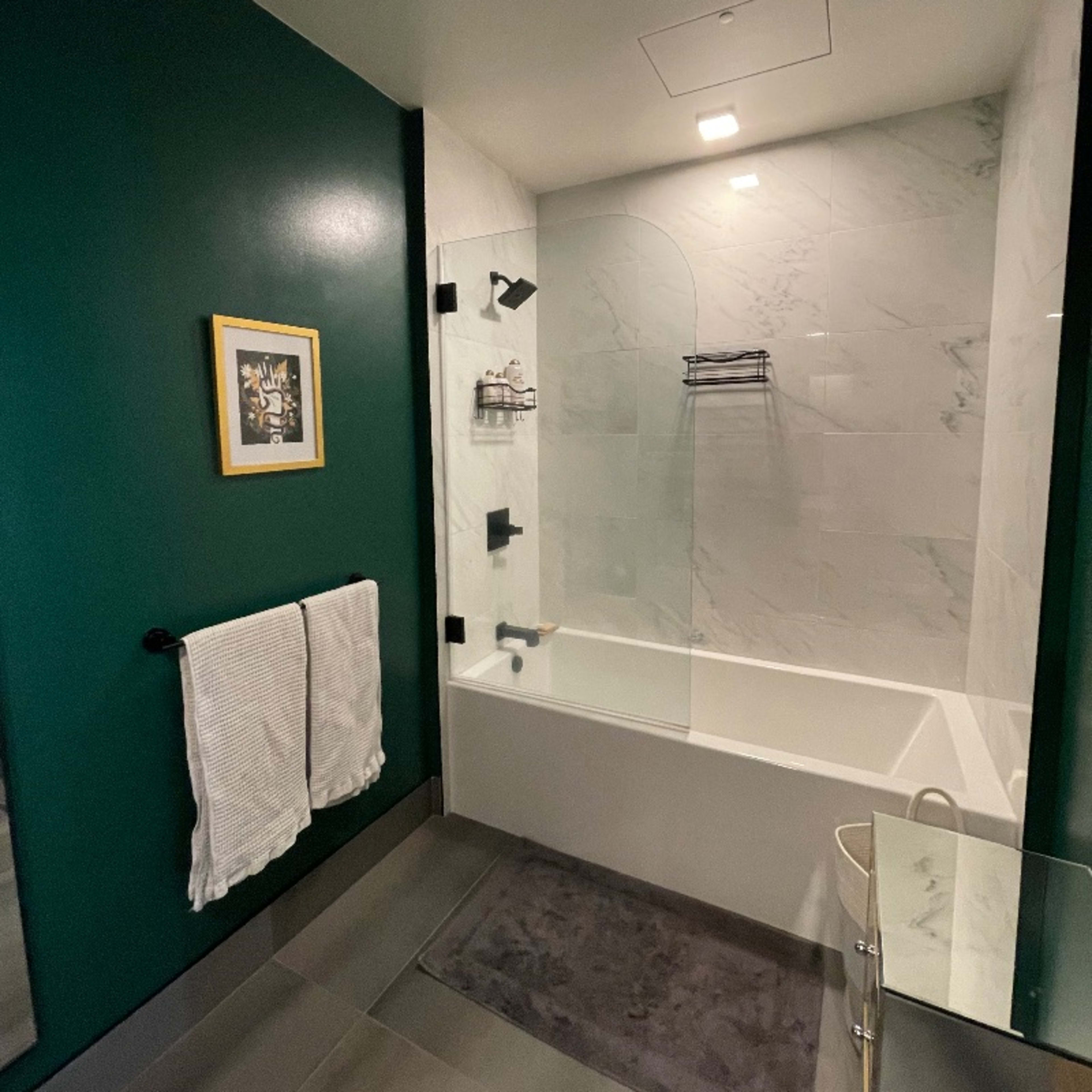 A bathroom with green walls, a white bathtub enclosed by a glass shower panel, and a framed picture on the wall.