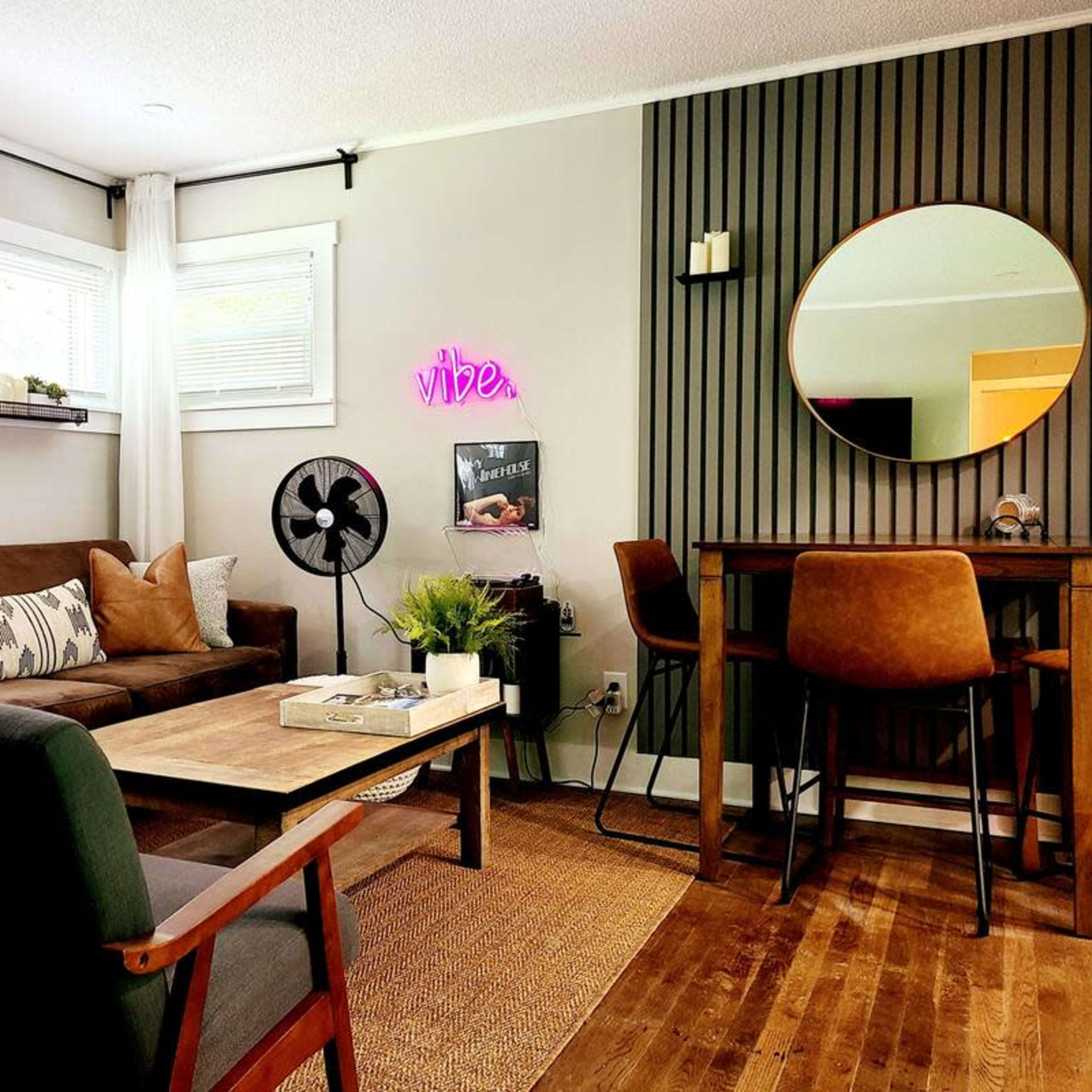 A modern living area with a brown sofa, two chairs, a circular mirror, and a small dining table near a wall with a neon sign.