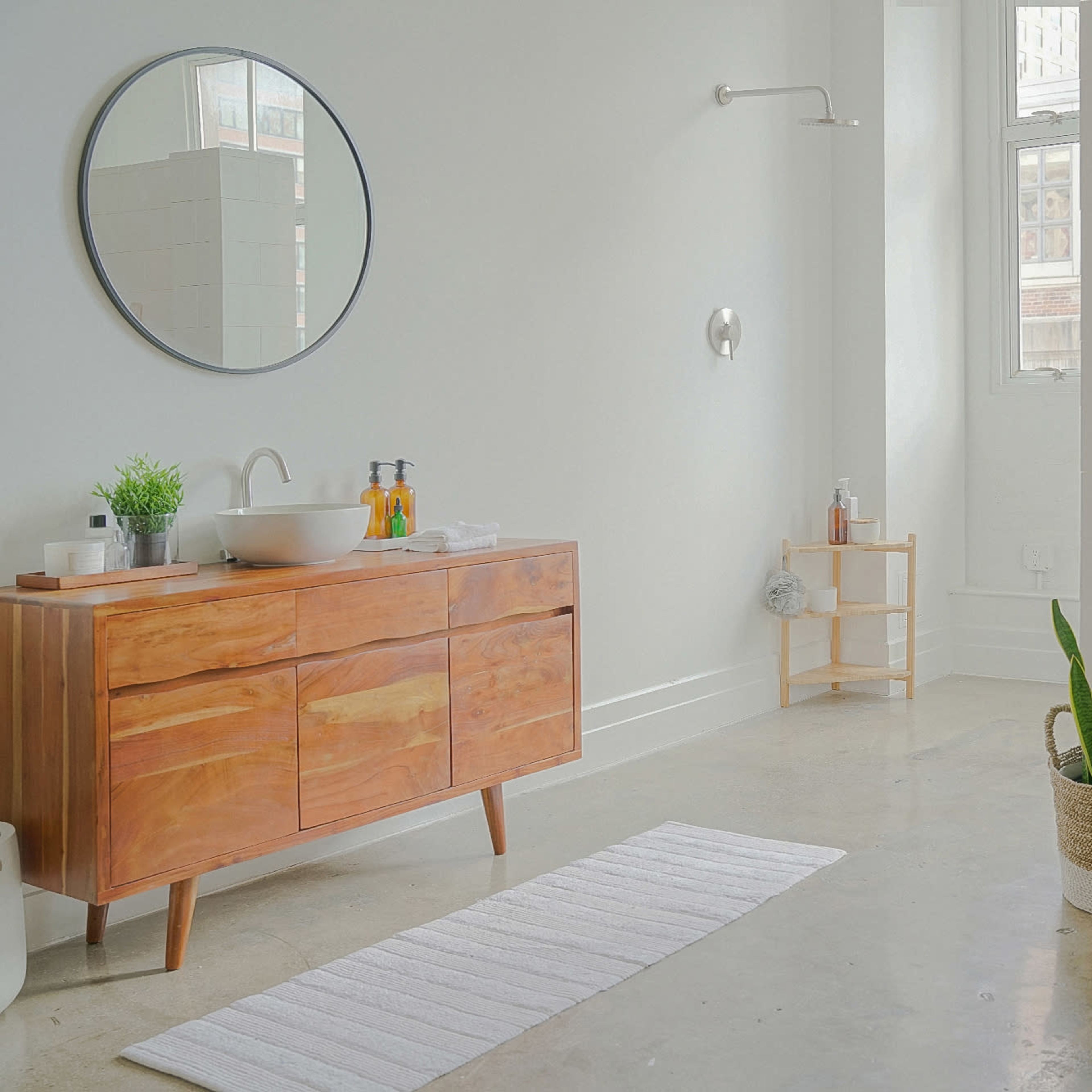 A modern bathroom features a wooden vanity with a round mirror, a shower area, a plant in a basket, and neatly arranged towels on a rack.