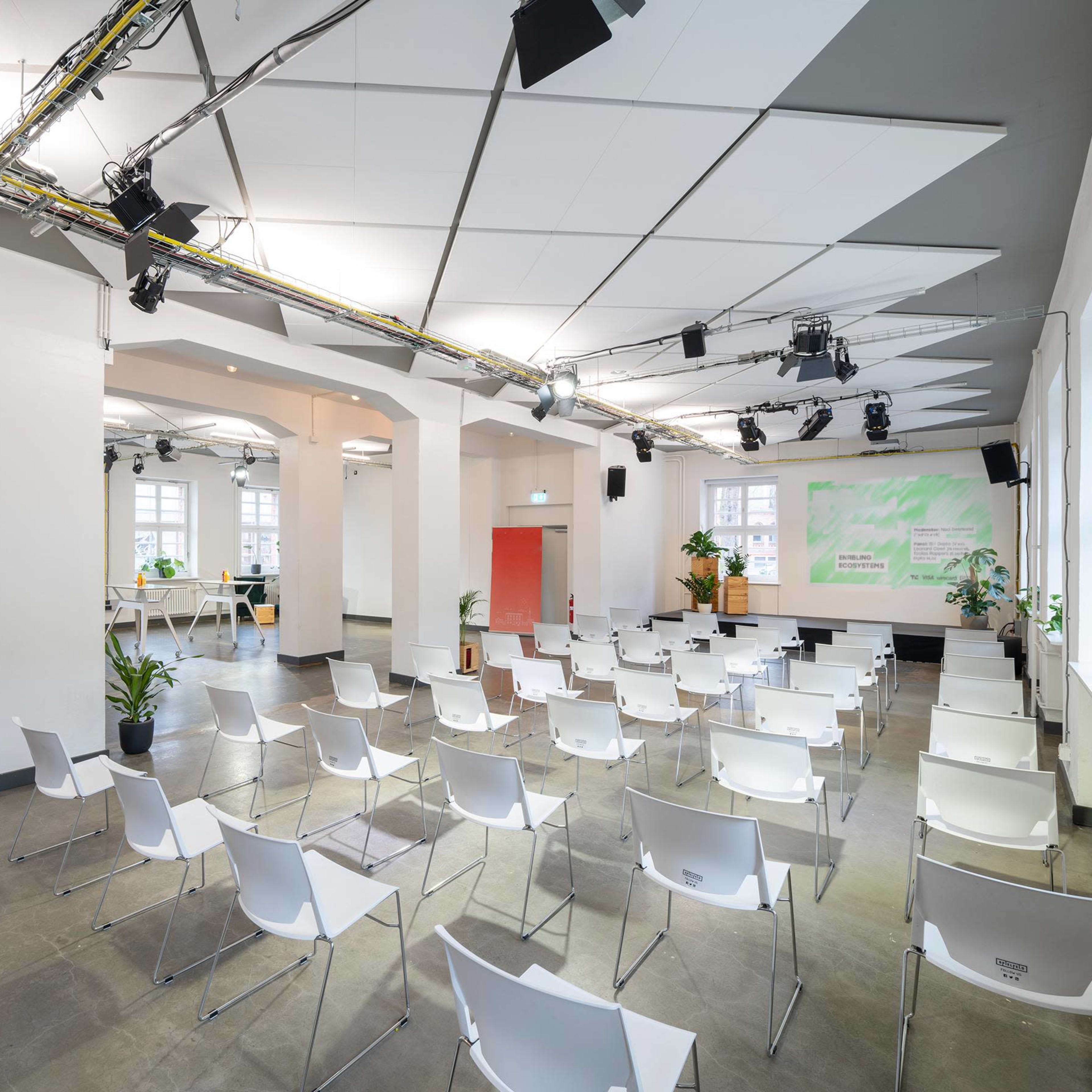 A spacious, modern conference room is set up with rows of white chairs facing a presentation area, featuring plants and bright lighting.