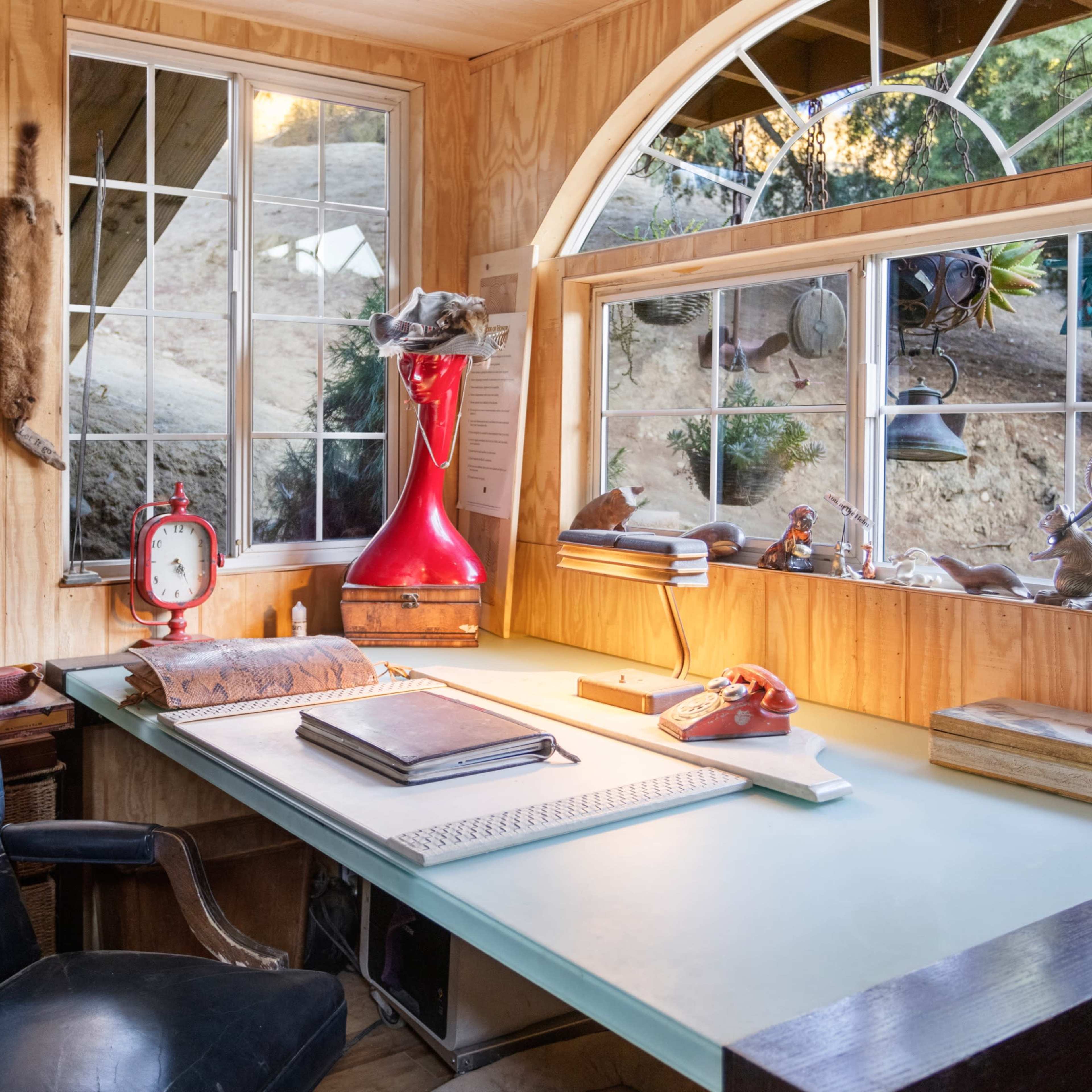 A wooden desk with a blue surface, an antique clock, and various items on it, positioned next to large windows with a view of greenery outside.