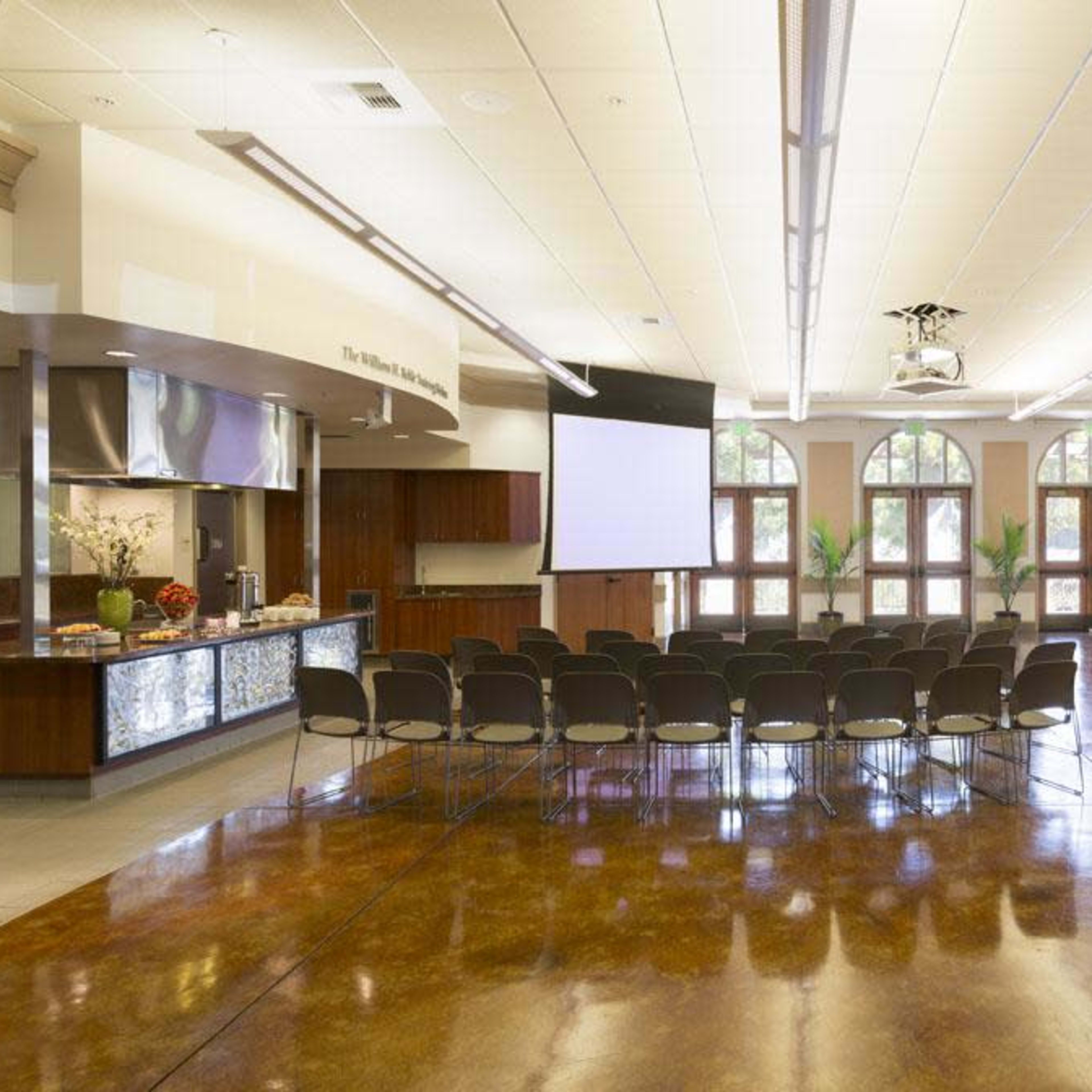 A spacious event room set up with rows of chairs facing a projection screen, with a refreshments table on one side.