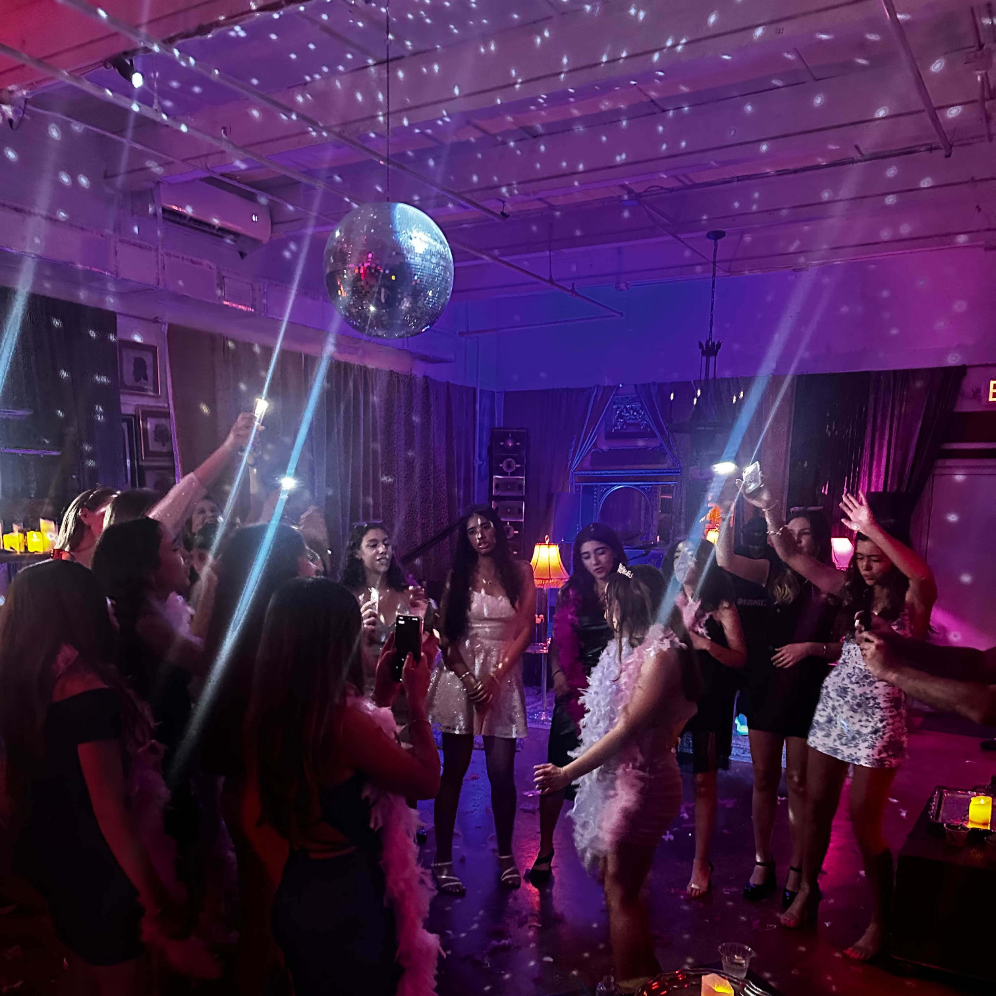A group of people celebrate in a dimly lit room adorned with a disco ball and colorful lights while holding drinks and dancing.