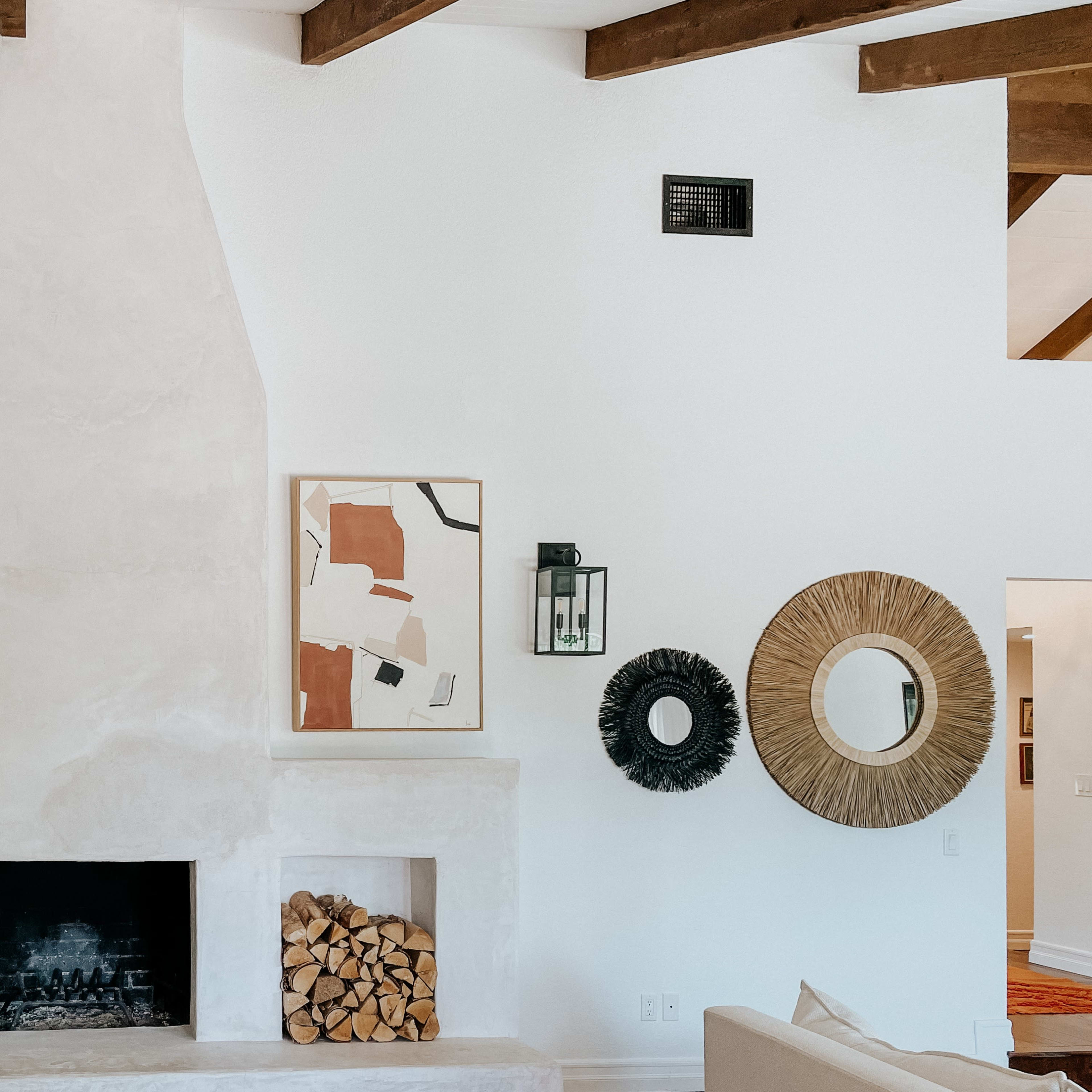 A living room featuring a white plaster fireplace, stacked firewood, decorative mirrors, and framed artwork on the wall, with wooden beams visible in the ceiling.