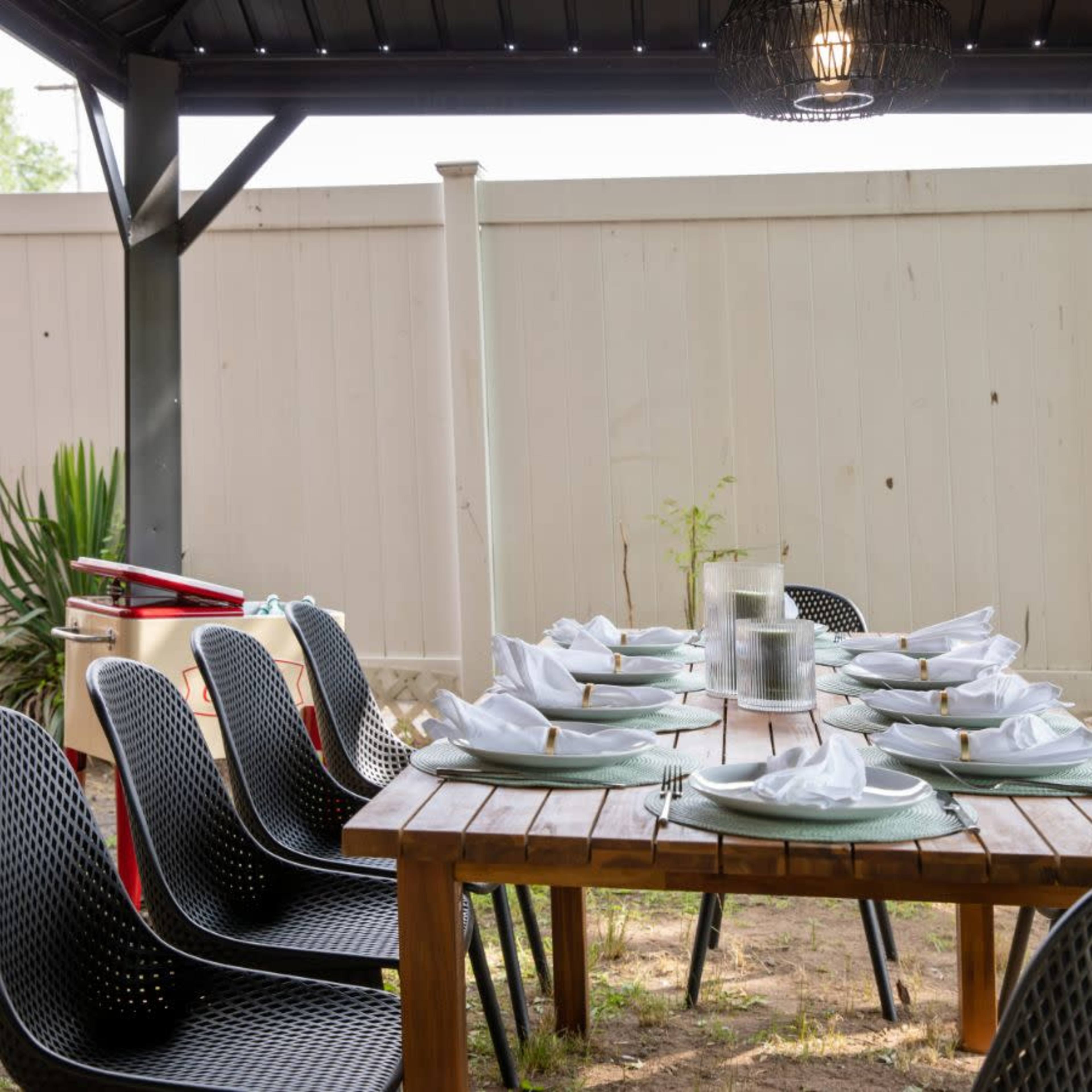 A long wooden table set with plates and napkins is arranged under a covered gazebo surrounded by black chairs and a fenced area.