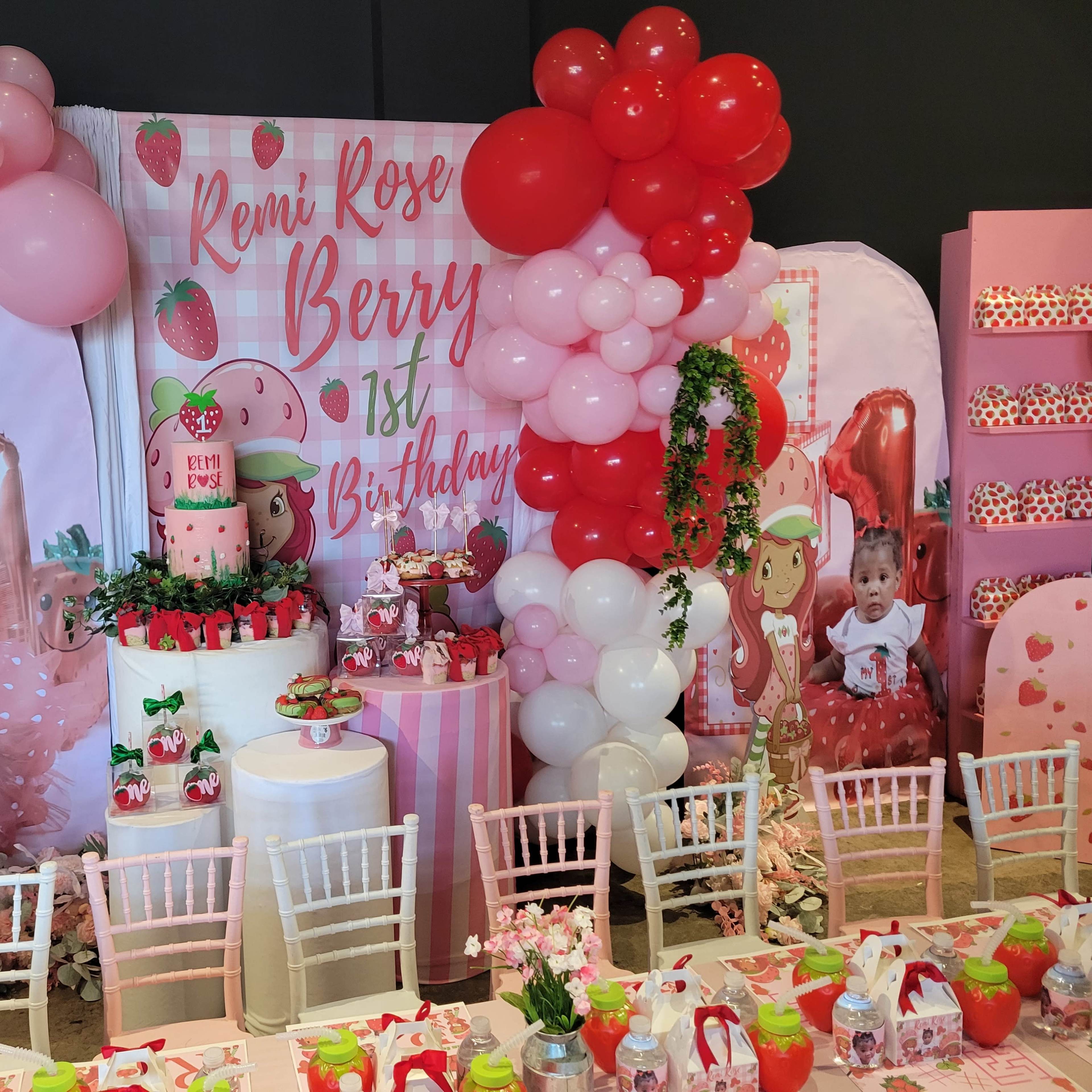 A colorful birthday party setup featuring a decorated backdrop with strawberries and balloons, a cake on a table, and themed decorations surrounding a dining area.