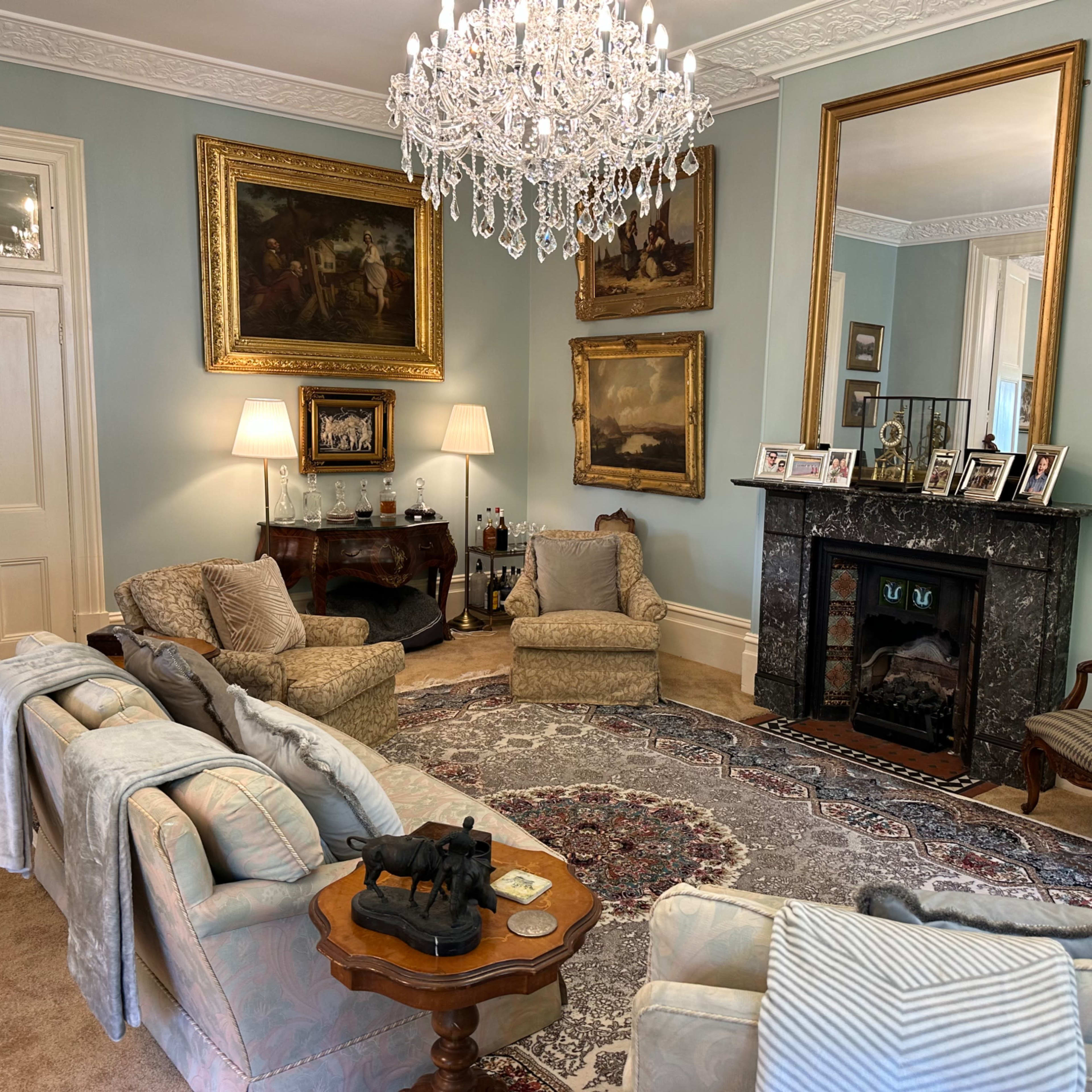 The living room features a large ornate chandelier, elegant seating, a marble fireplace, and framed artwork on the walls.