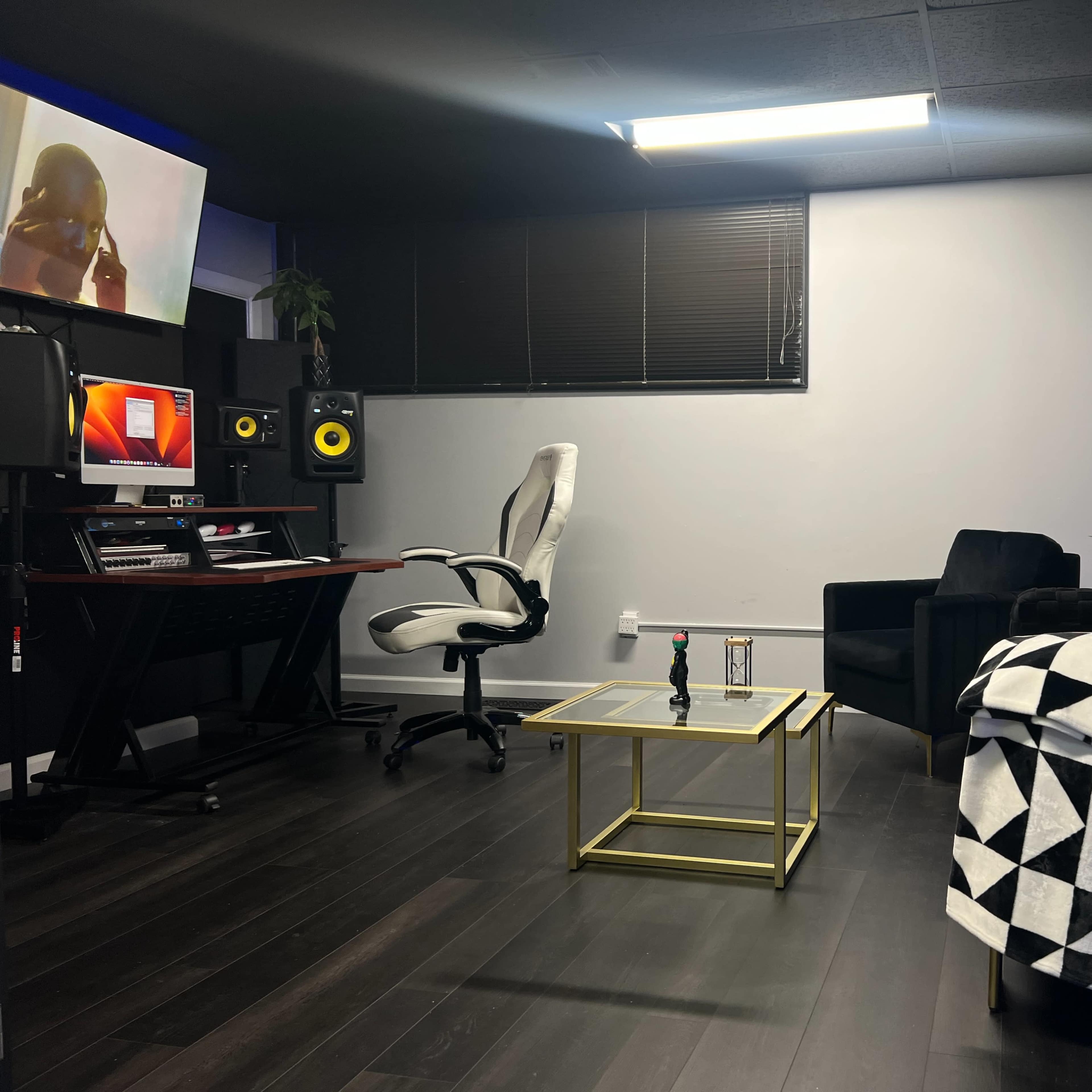 A modern recording studio with a desk, computer, sound monitors, a comfortable chair, and a black couch, all set against dark flooring and gray acoustic panels.