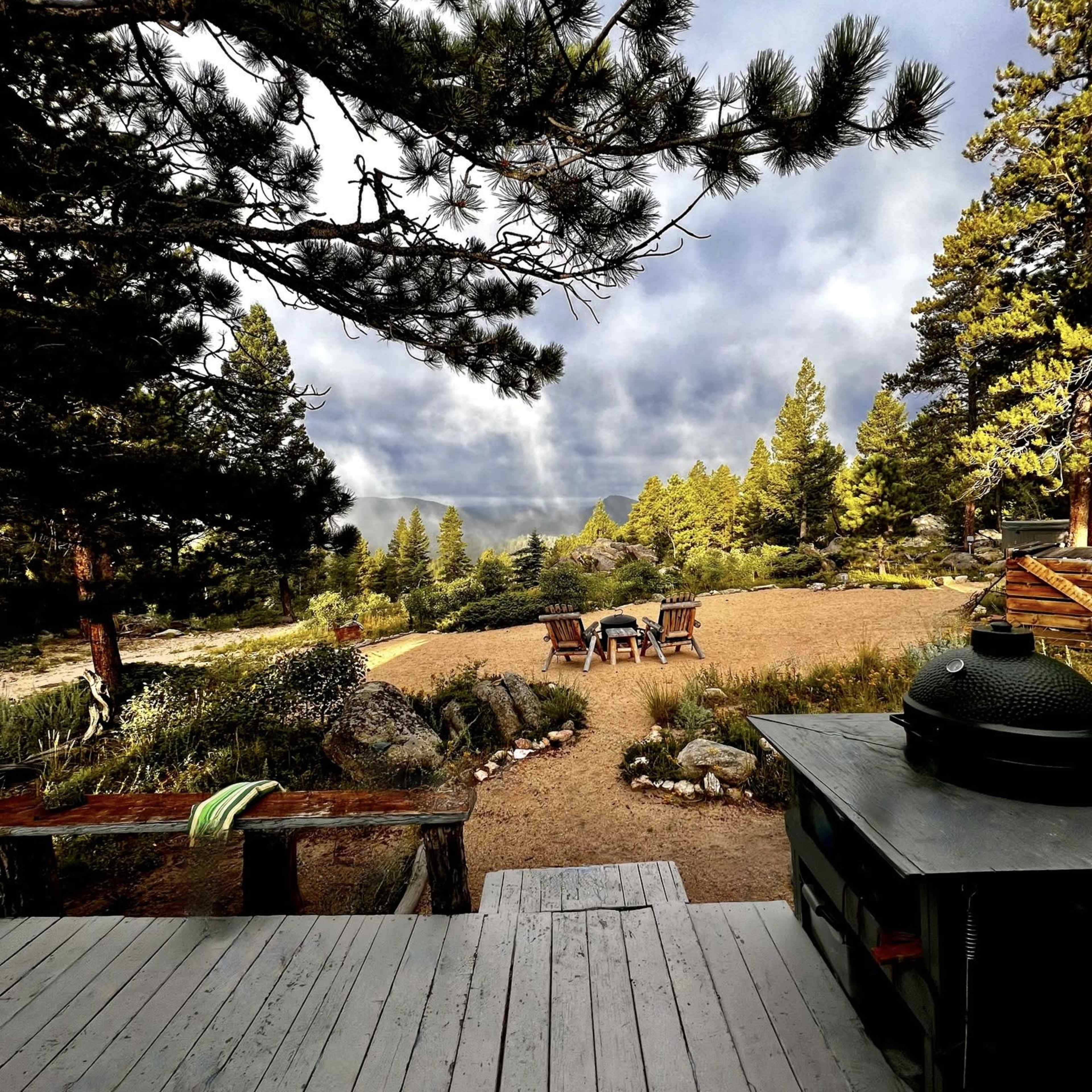 A wooden deck overlooks a landscaped area with a fire pit surrounded by chairs, framed by trees and distant hills under a cloudy sky.
