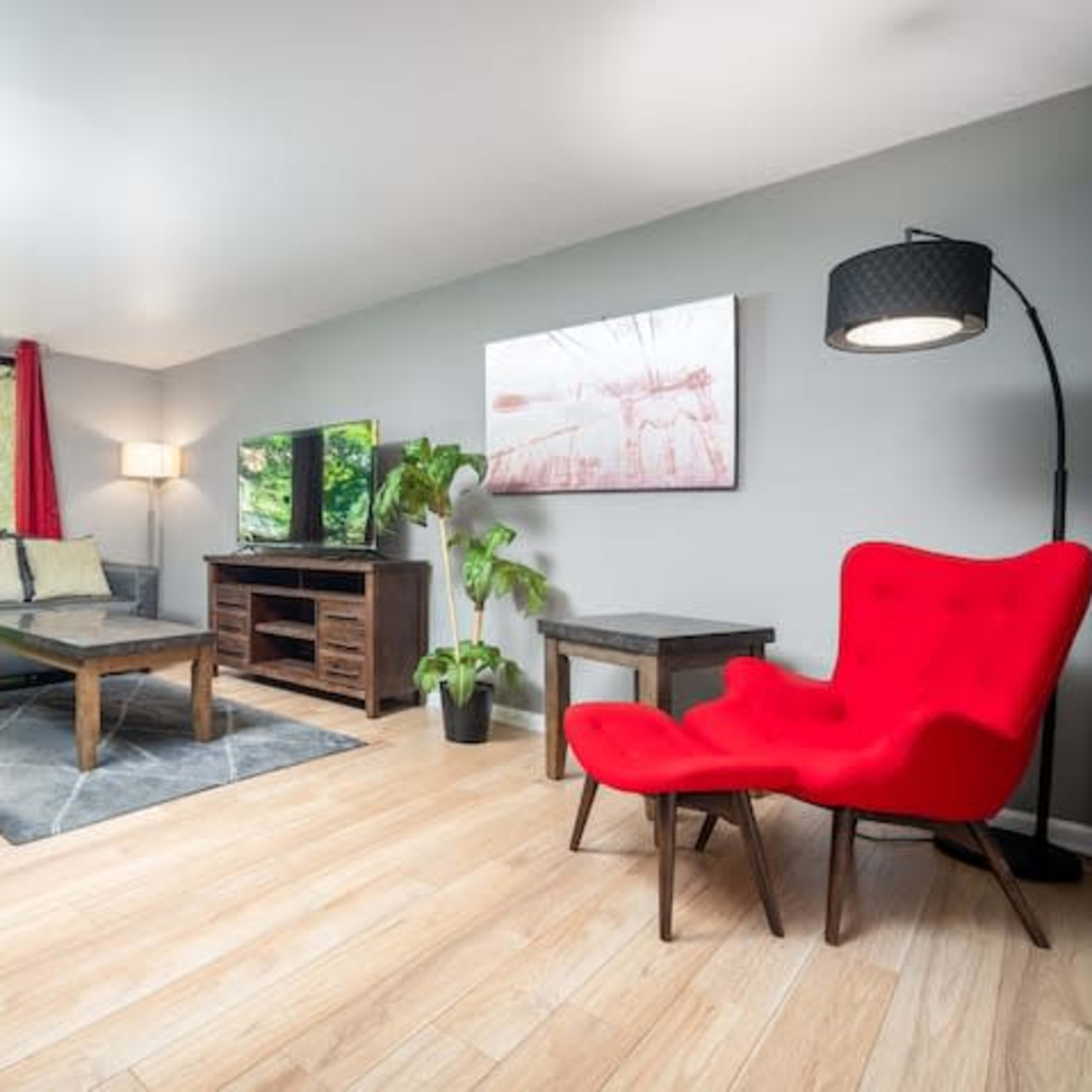A modern living room with a gray sofa, a red accent chair, a wooden coffee table, and a television, framed by large windows with red curtains overlooking a green outdoor area.