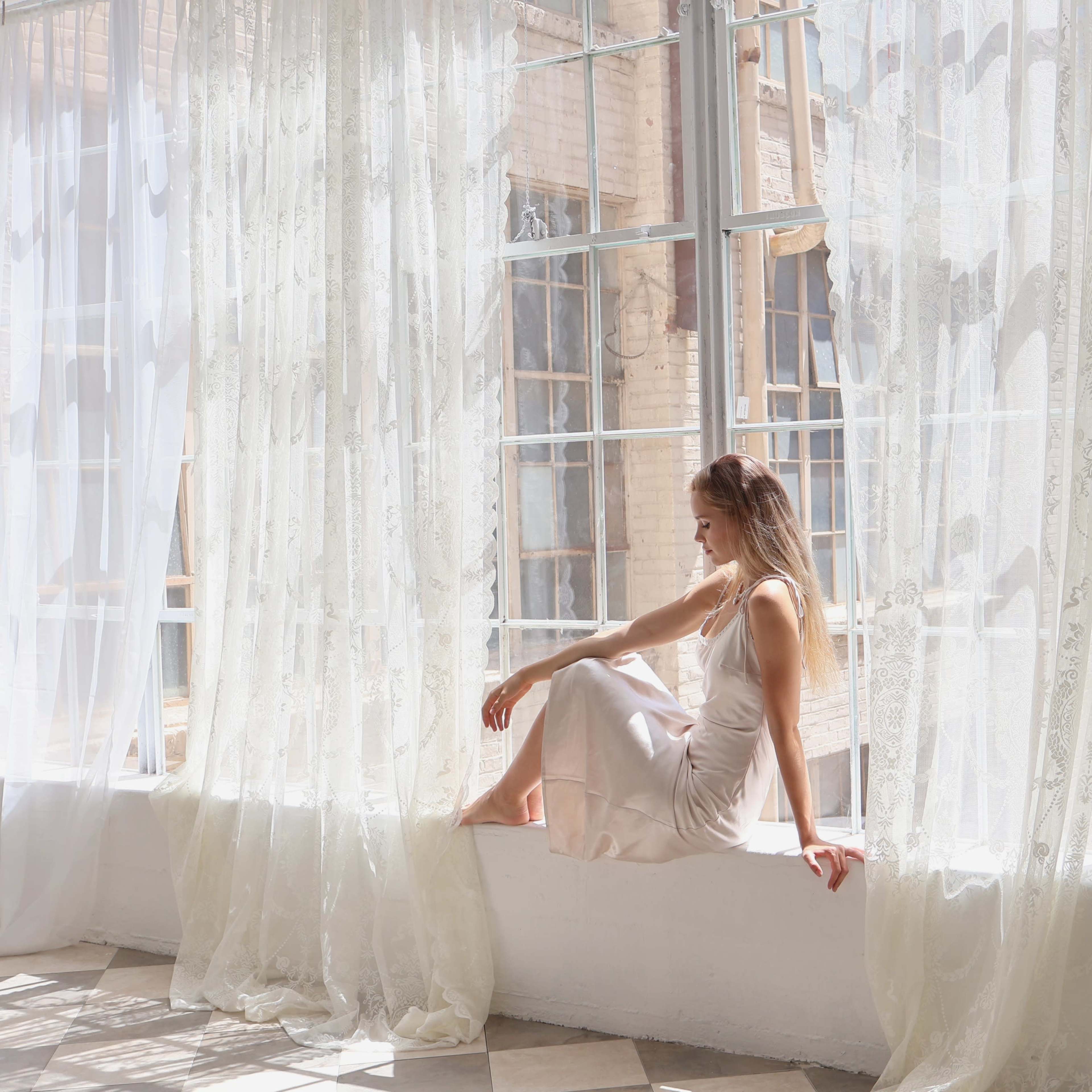 A woman in a light dress sits on a windowsill, surrounded by flowing white curtains and bright natural light.