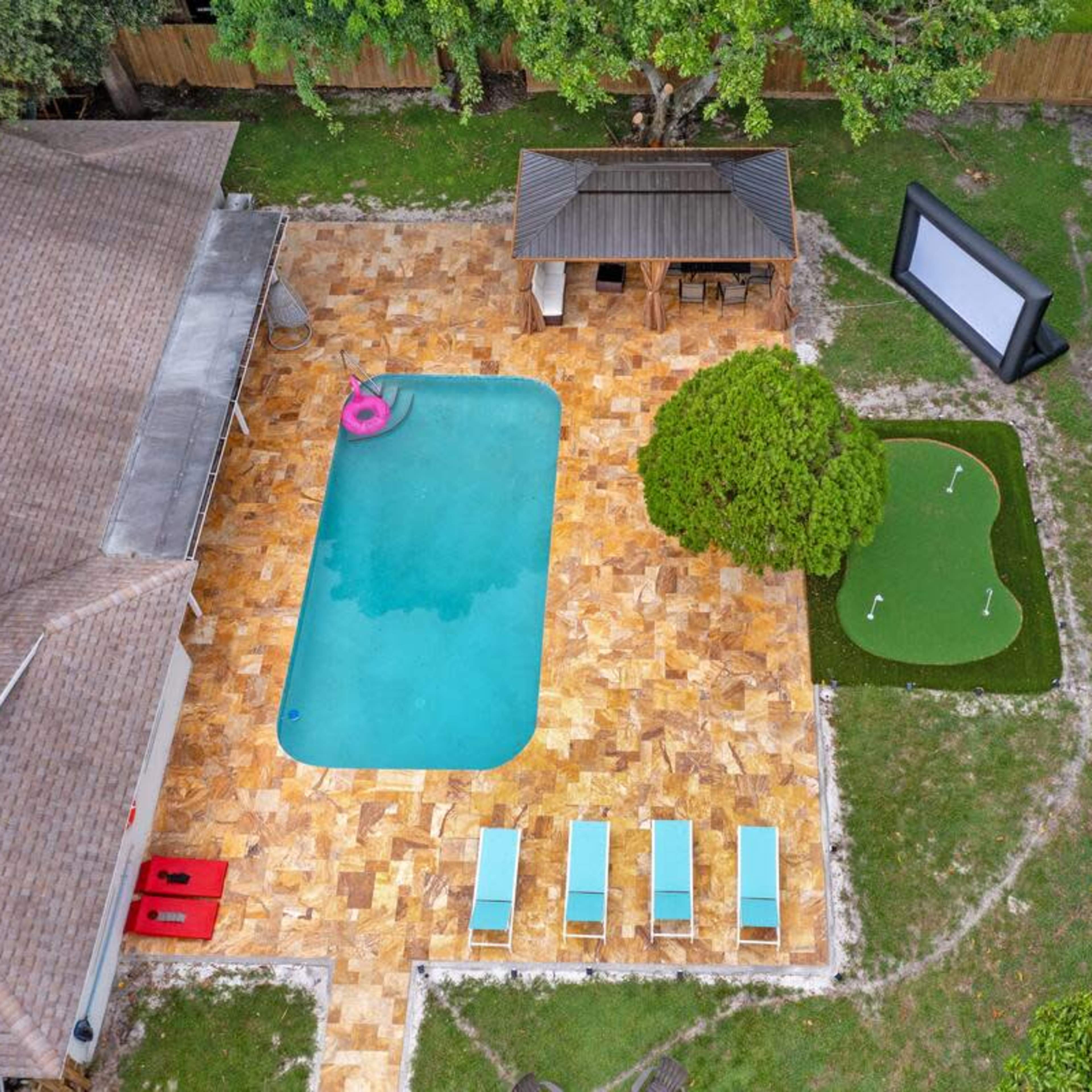 An aerial view of a residential backyard featuring a rectangular swimming pool, lounge chairs, a gazebo, and a small putting green adjacent to a large screen.