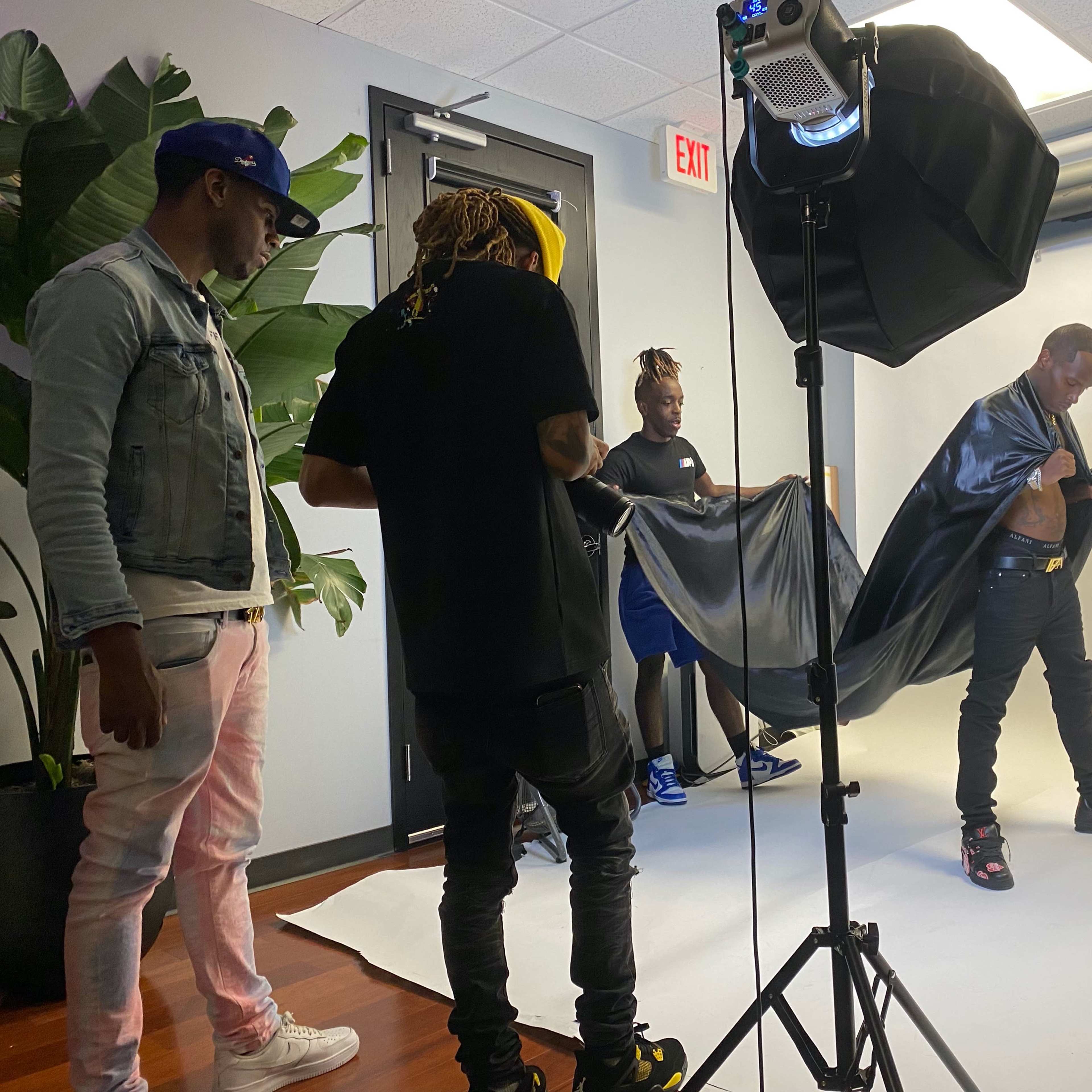 A group of four people are in a photography studio, with one person adjusting their outfit in front of a backdrop while others assist.