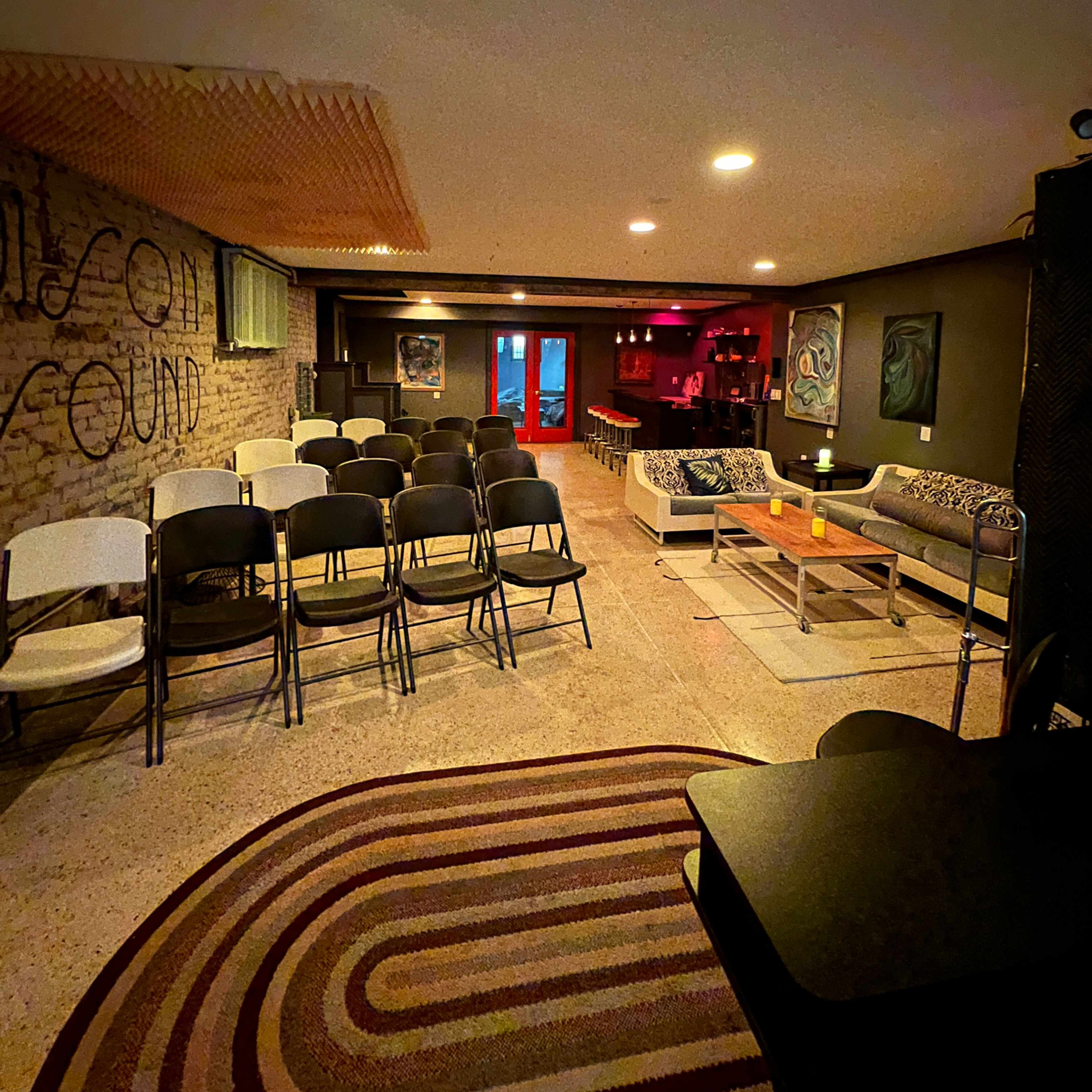 A dimly lit lounge area with folding chairs arranged in rows, a bar in the background, and comfortable seating nearby.