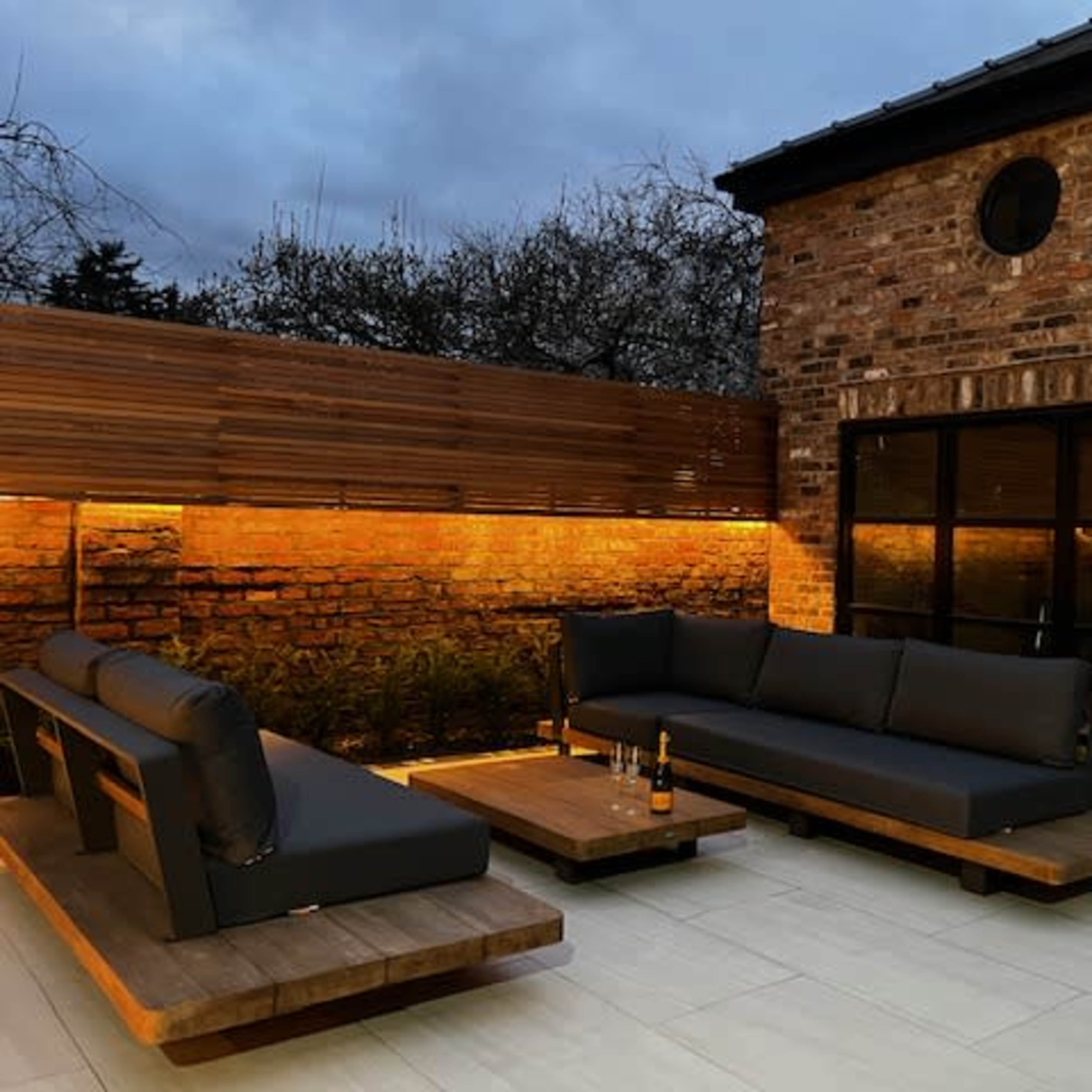 A modern outdoor seating area features dark upholstered sofas and wooden coffee tables against a textured brick wall with ambient lighting.